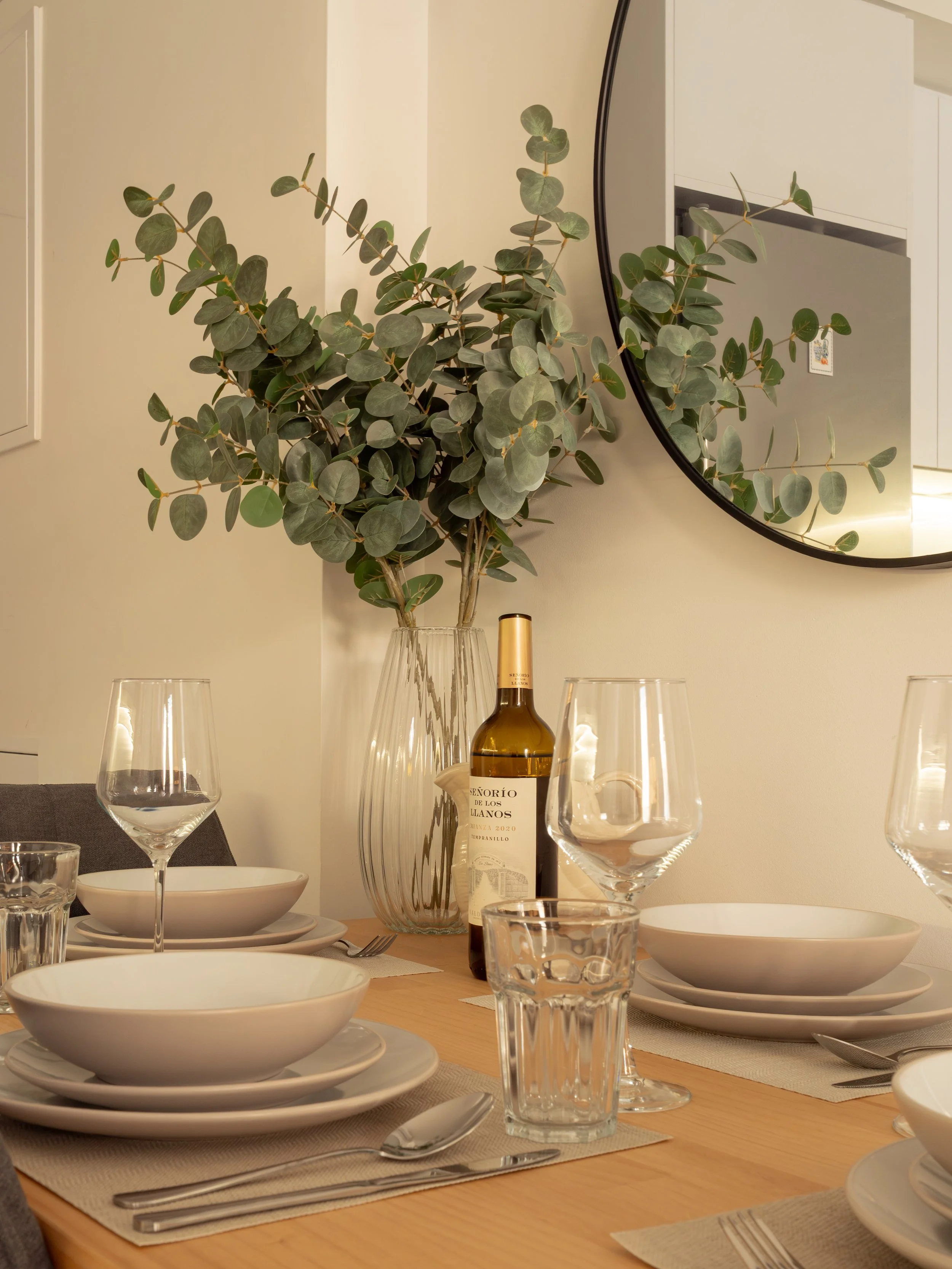 A dining table set with white bowls, drinking glasses, silverware on beige placemats, a bottle of white wine, and a large mirror reflecting a green plant and kitchen cabinets behind it.