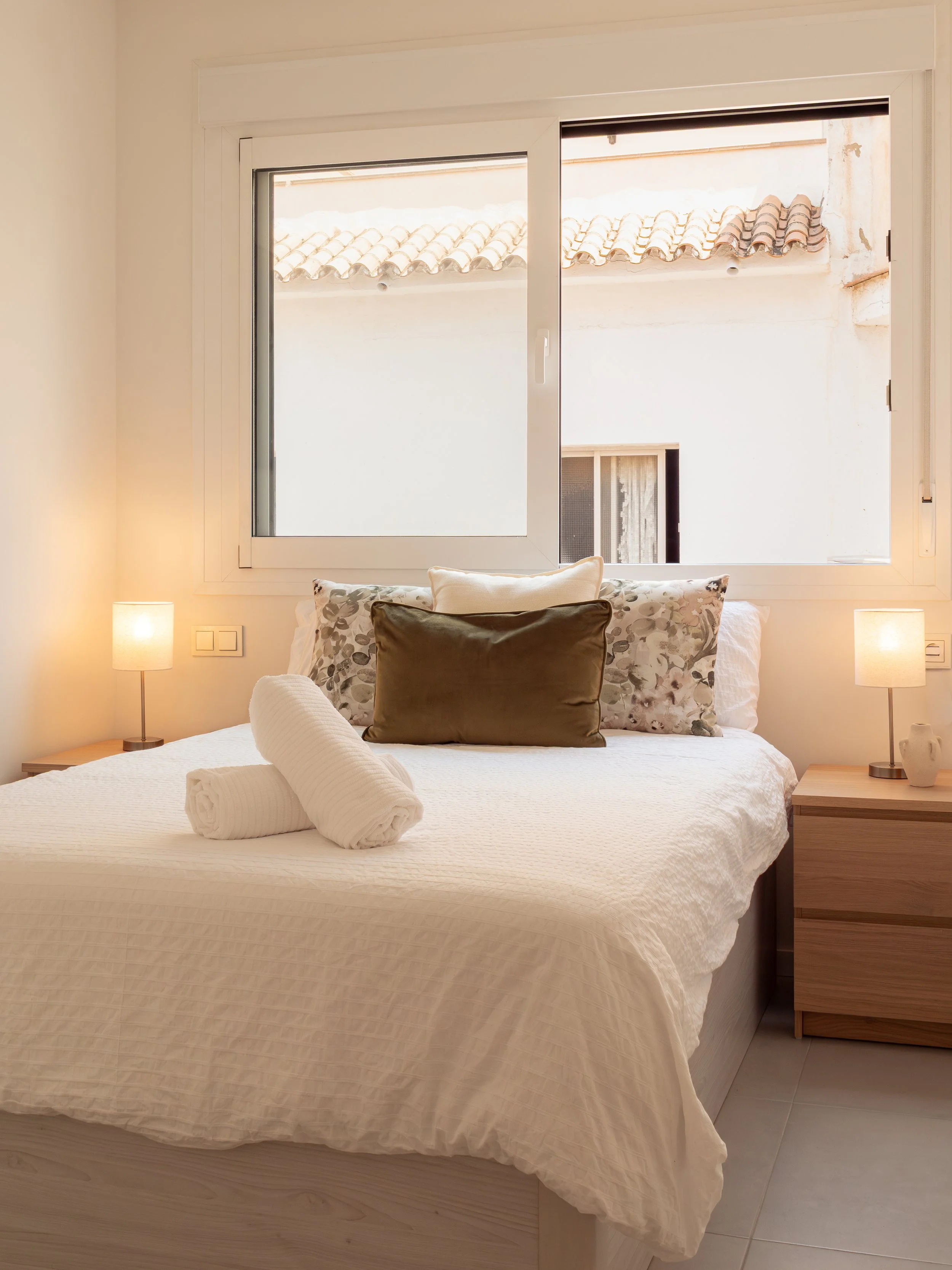 A bedroom with a bed covered in white bedding, centered in front of a large window showing a neighboring building with tiled roof, two lamps on bedside tables, and pillows including a rolled towel on the bed.