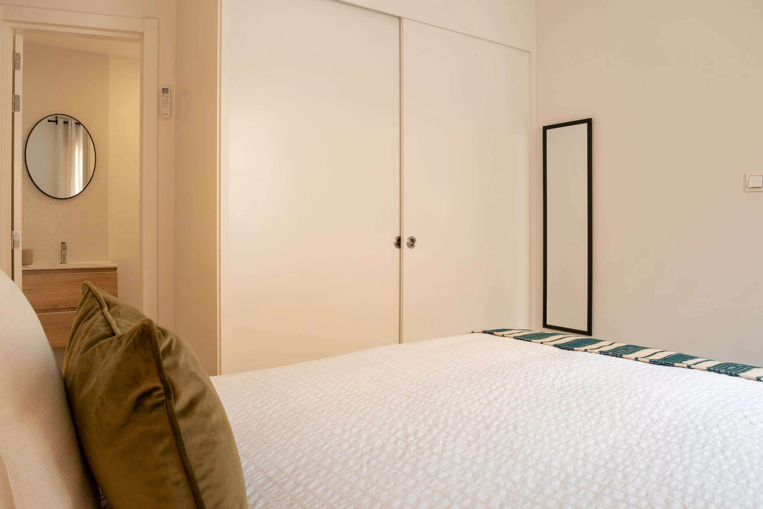 A bedroom with a white bedspread, tan pillows, a large mirror on a black frame leaning against the wall, a white closet with sliding doors, a black and white striped blanket on the bed, and a bathroom with a round mirror and sink visible through an o