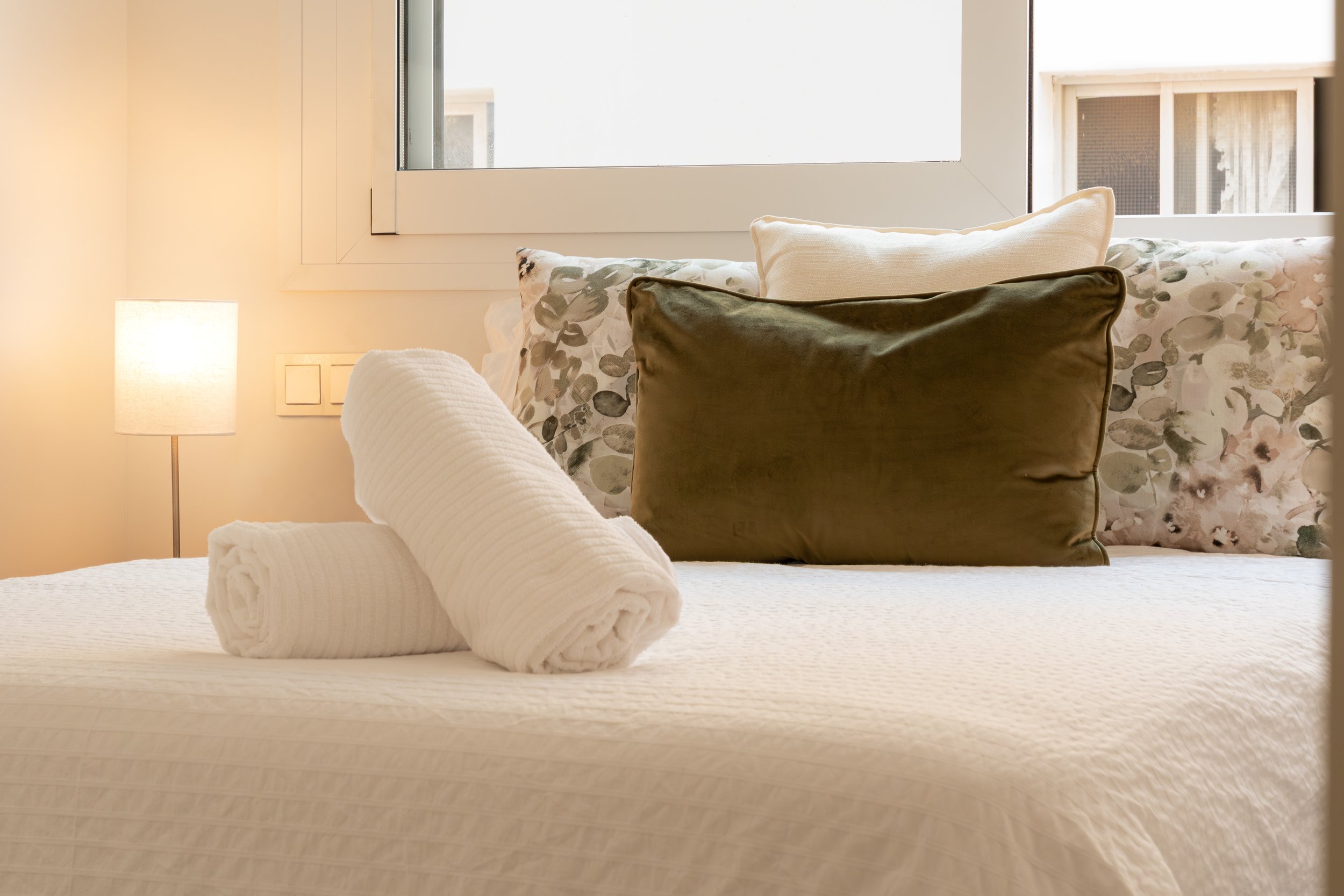 A neatly made bed with rolled white towels at the foot, floral and dark green pillows, and a white headboard. There is a window with white framing, a small lamp, and beige walls in the room.