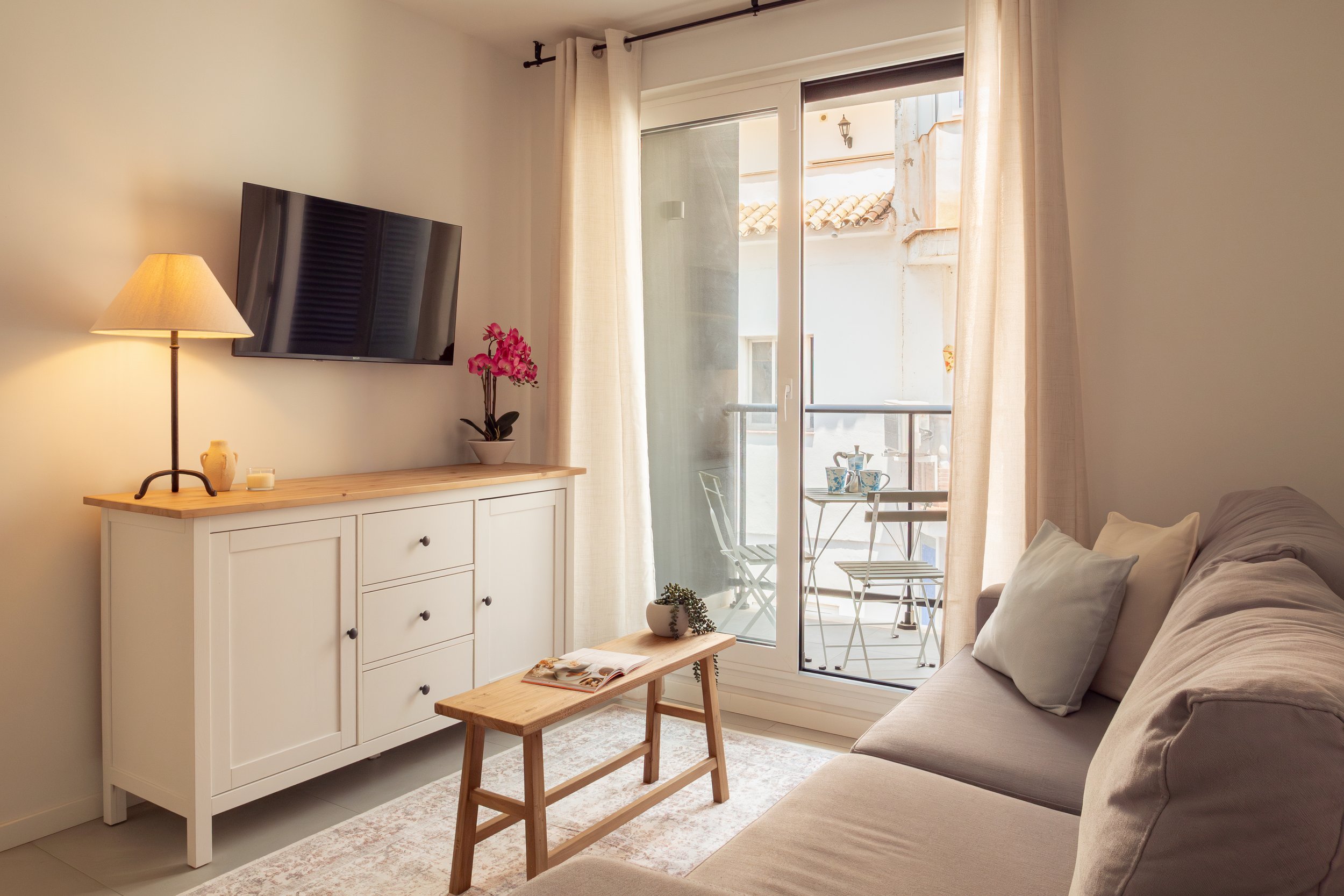 A cozy living room with a beige sofa, a white cabinet with drawers, a wooden coffee table, a lamp, a potted pink orchid, a large flat-screen TV on the wall, and a balcony with a small table and chairs, all illuminated by natural light through a slidi