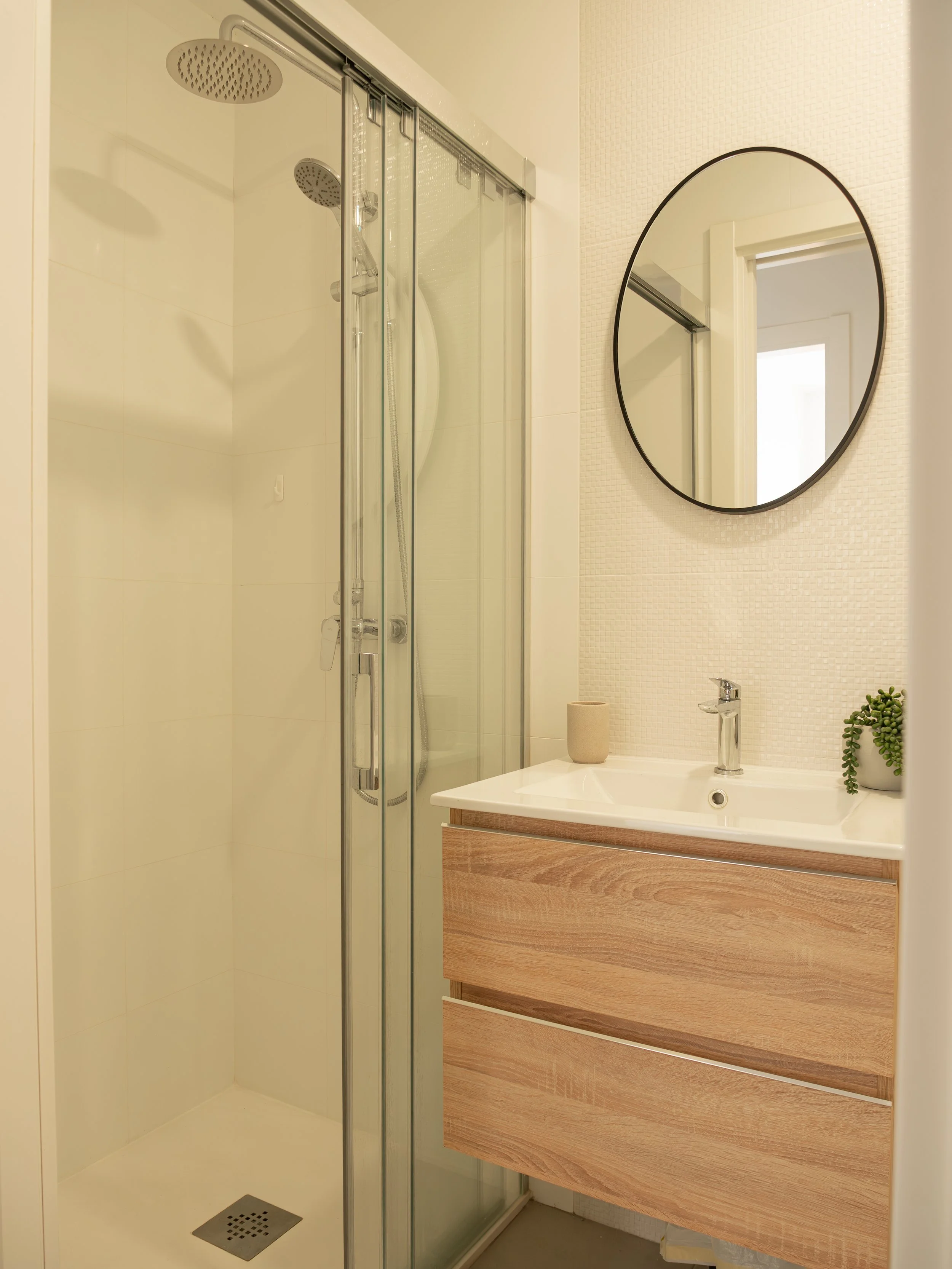 A modern bathroom featuring a glass-enclosed shower with two showerheads, a large round mirror, a wooden vanity with a white sink and a silver faucet, a small plant, a soap dispenser, and a beige textured wall.