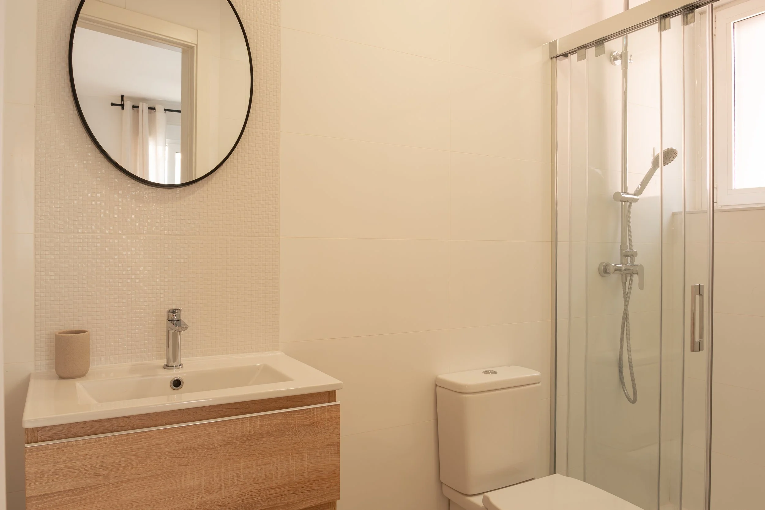 A clean, modern bathroom with a mirror, sink, toilet, and a shower stall with glass doors. The bathroom features white tiled walls and a window bringing in natural light.
