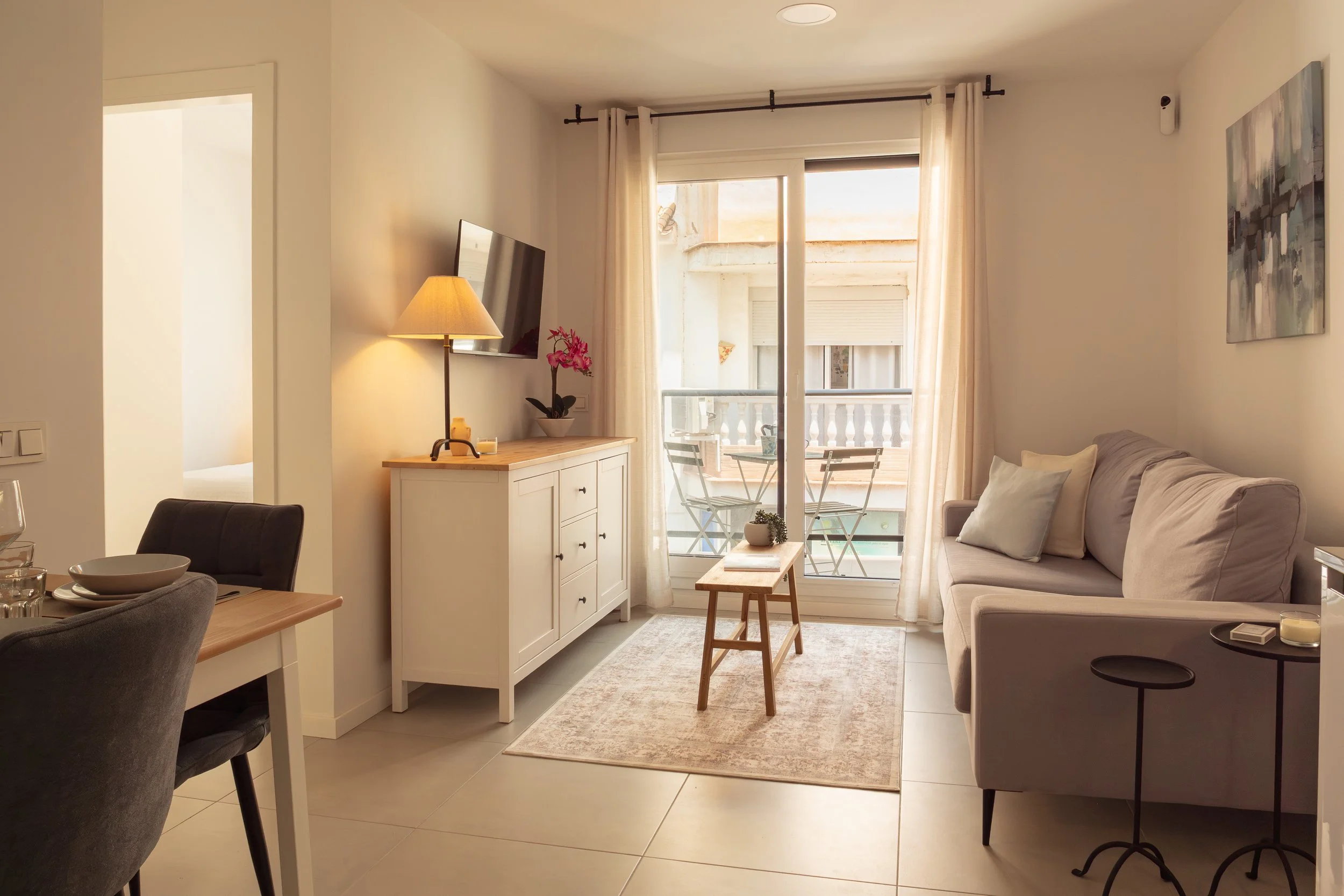 Living room with beige couch, small coffee table, and sliding glass door with curtains leading to a balcony with outdoor chairs and a railing. There's a white sideboard with a pink orchid and wall-mounted TV, with artwork on the wall and a lamp on th