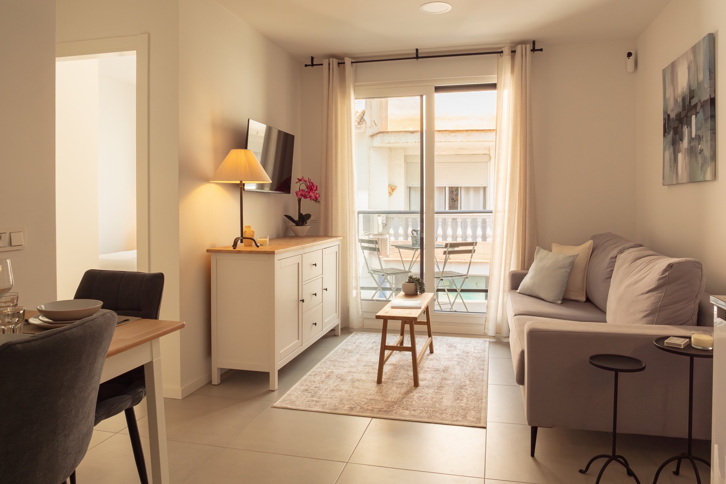 A cozy living room with a beige sofa and pillows, a white cabinet with a pink orchid, a wall-mounted TV, a floor lamp, a small wooden table, a patio with two chairs and a small table outside, large glass doors with light curtains, artwork on the wall