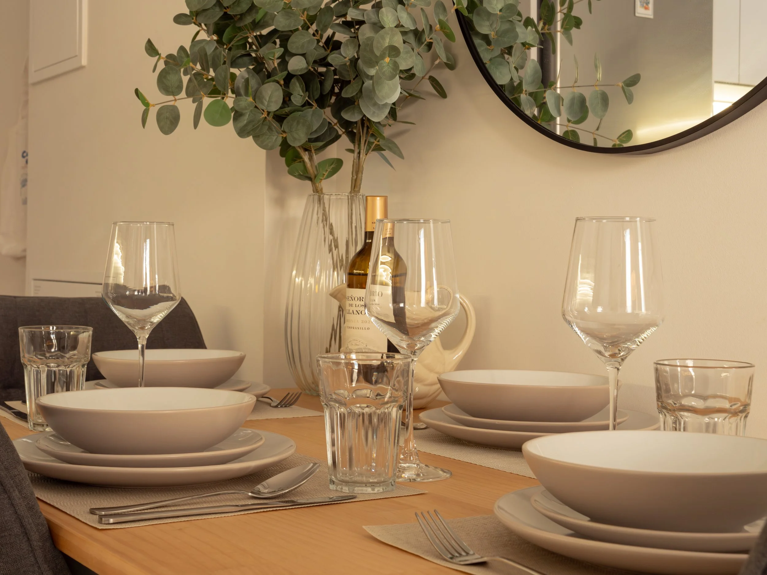 A dining table set with white bowls, empty wine glasses, water glasses, white plates, silverware, and a beige table runner. A large green leafy plant in a glass vase and a wine bottle are in the background, along with a round mirror on the wall refle