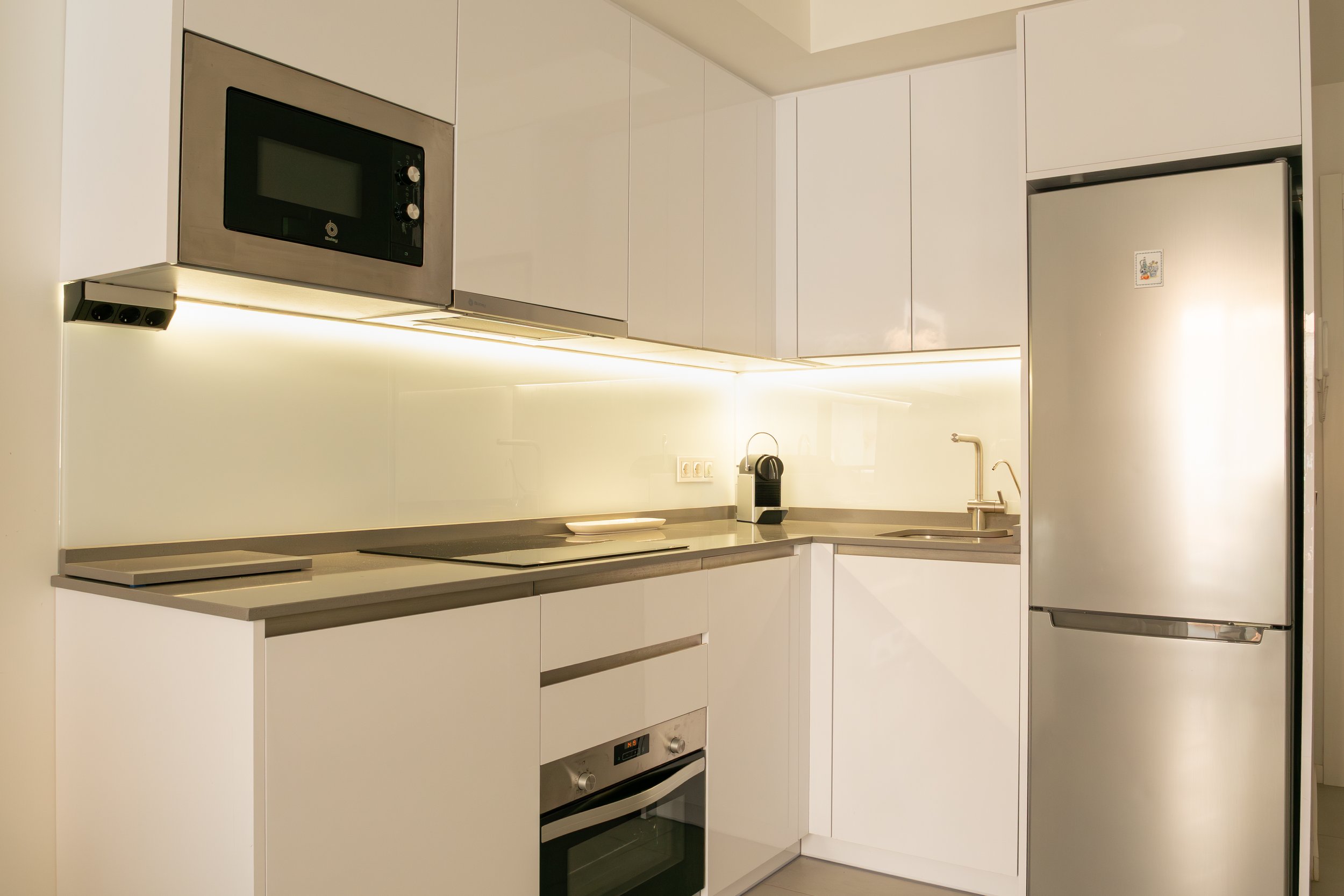 A modern minimalist kitchen with white cabinets, a built-in microwave, an oven, and a stainless steel refrigerator. The kitchen has a clean, sleek design with under-cabinet lighting and a small countertop with a coffee machine and a plate.