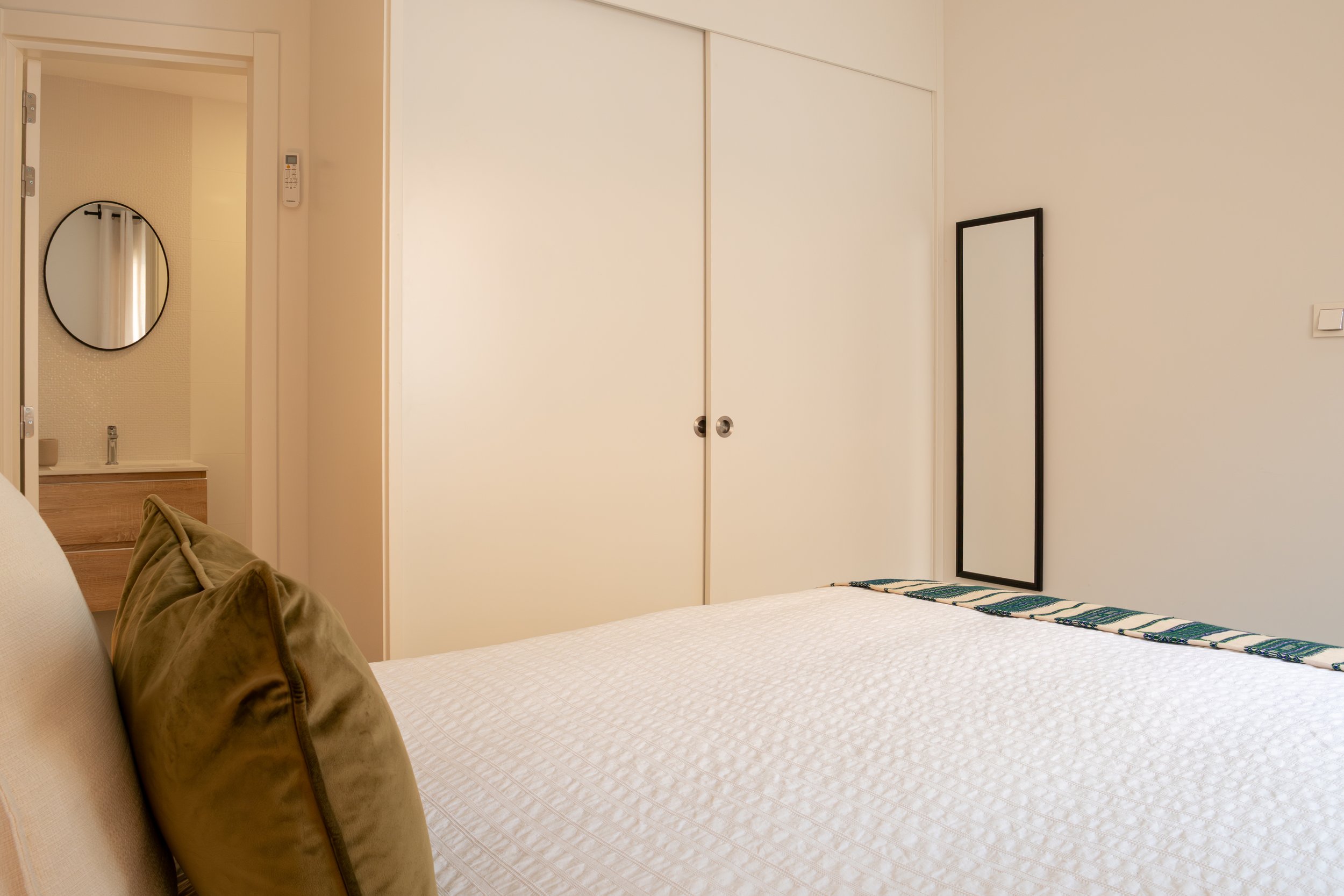 A neatly made bed with a white quilt and a patterned throw, a brown velvet pillow, and a mirror leaning against the wall. There is a sliding door wardrobe and a framed mirror on the right side of the room. A small side table with a sink and a round m