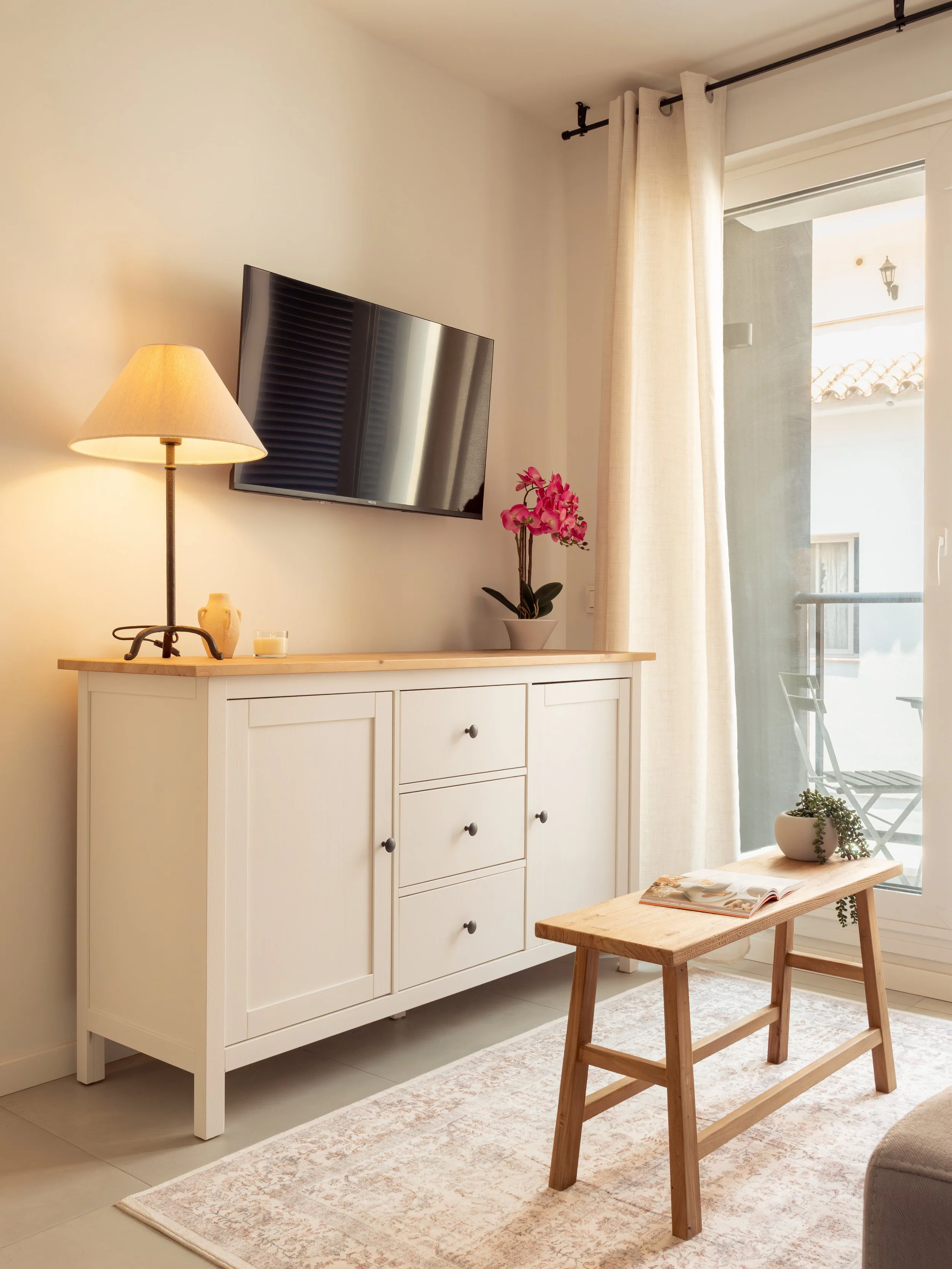 Living room corner with a white cabinet, a standing lamp, a TV, a pink orchid in a pot, a small wooden stool with an open book and small potted plant, beige curtains, and a balcony door with outdoor furniture visible.