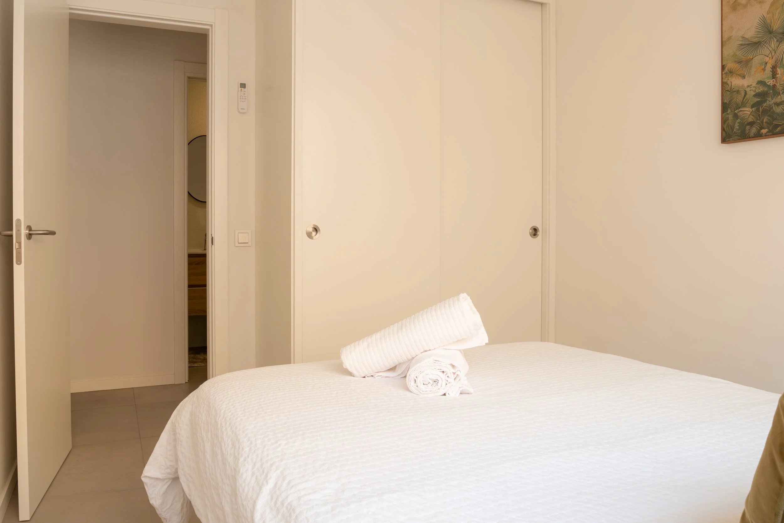 A bedroom with a bed covered in a white textured bedspread, two rolled towels on the bed, and beige walls with sliding closet doors and a small picture on the right wall.