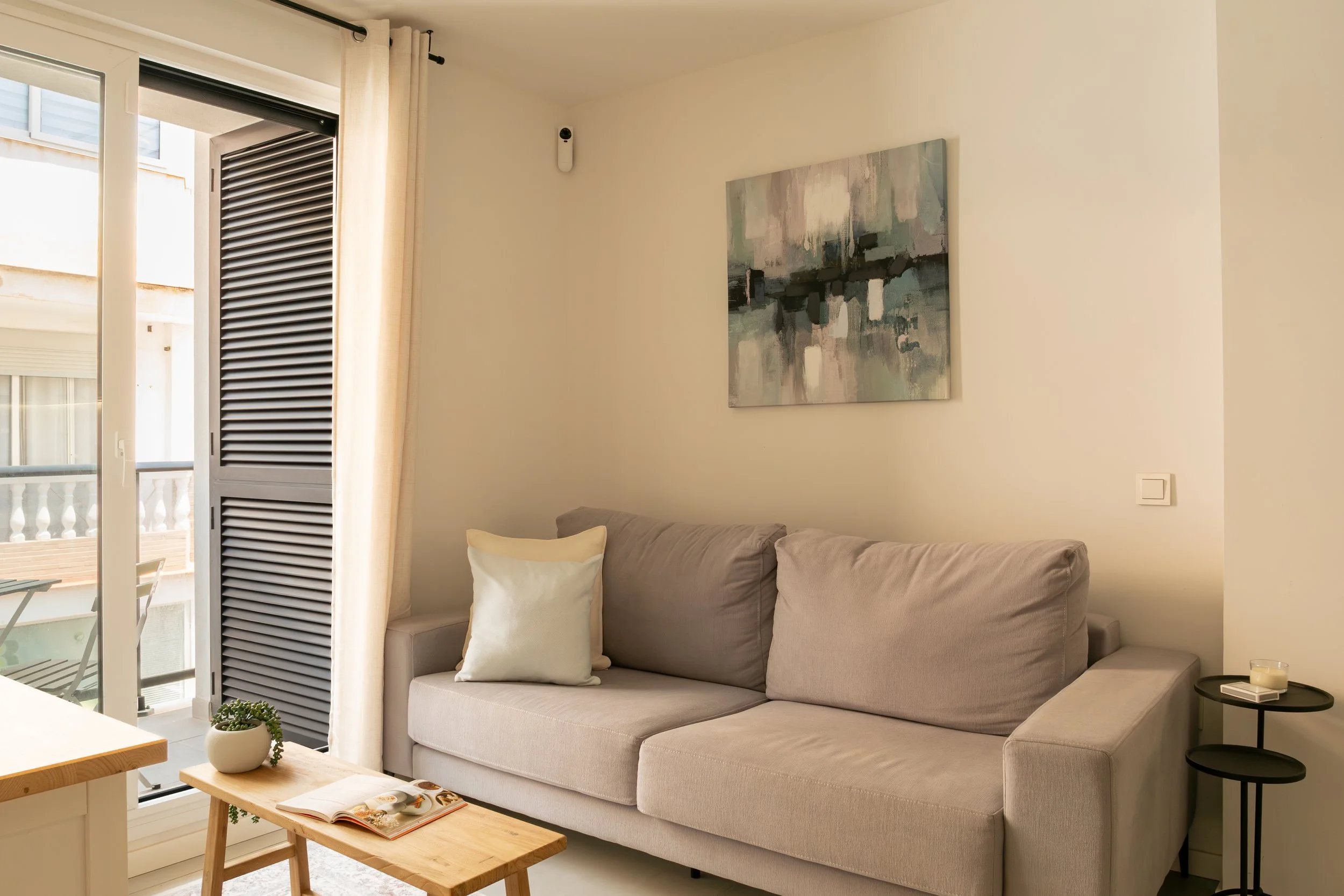A bright living room with a beige sofa, a white cushion, a small wooden coffee table with an open book, a small plant, and a black side table with a candle. There's a large window with white curtains, a balcony outside, and a wall-mounted device near