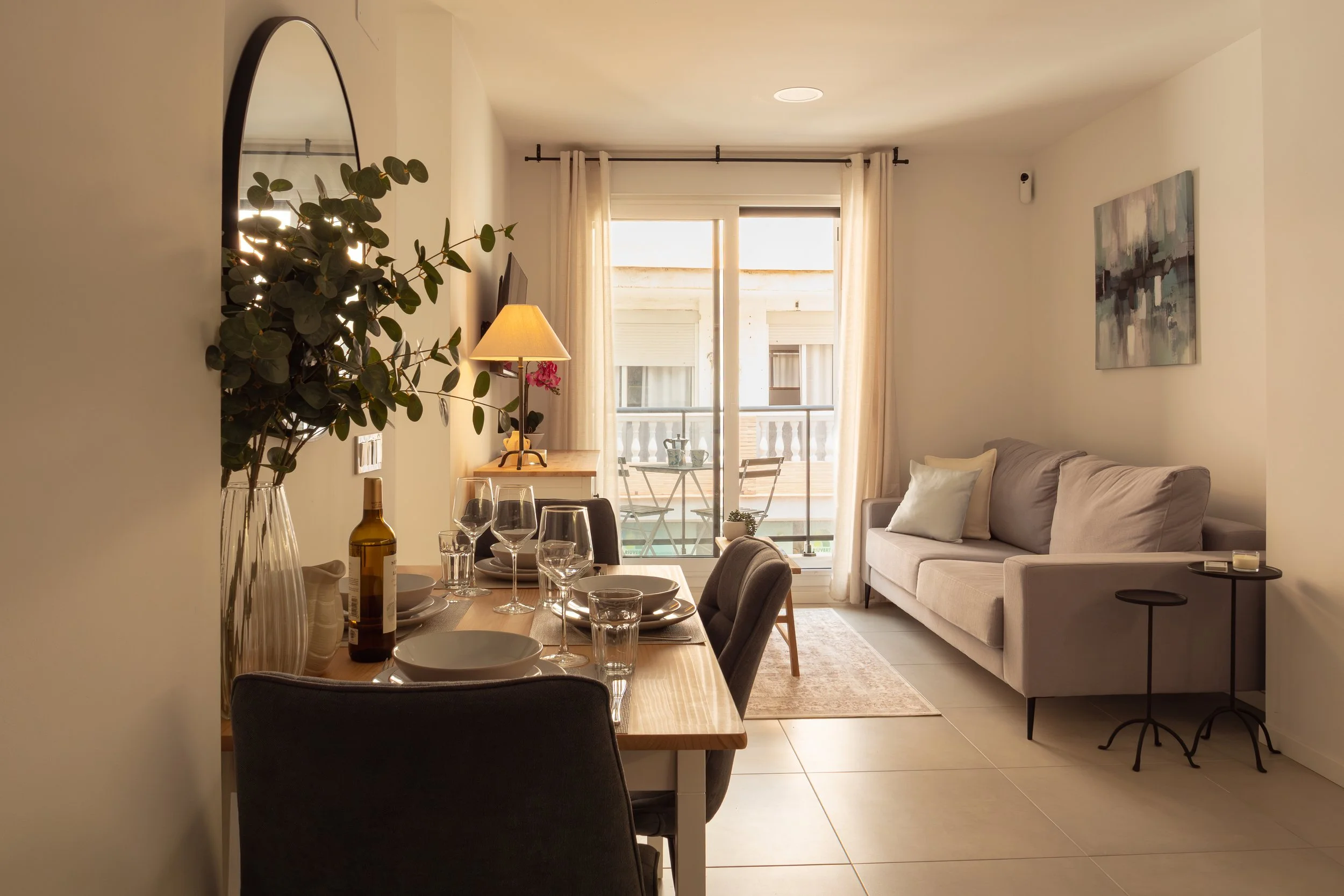 Living room with beige sofa, wall art, sliding glass door with white curtains, dining table set with glasses and plates, indoor plant, and a balcony visible outside.