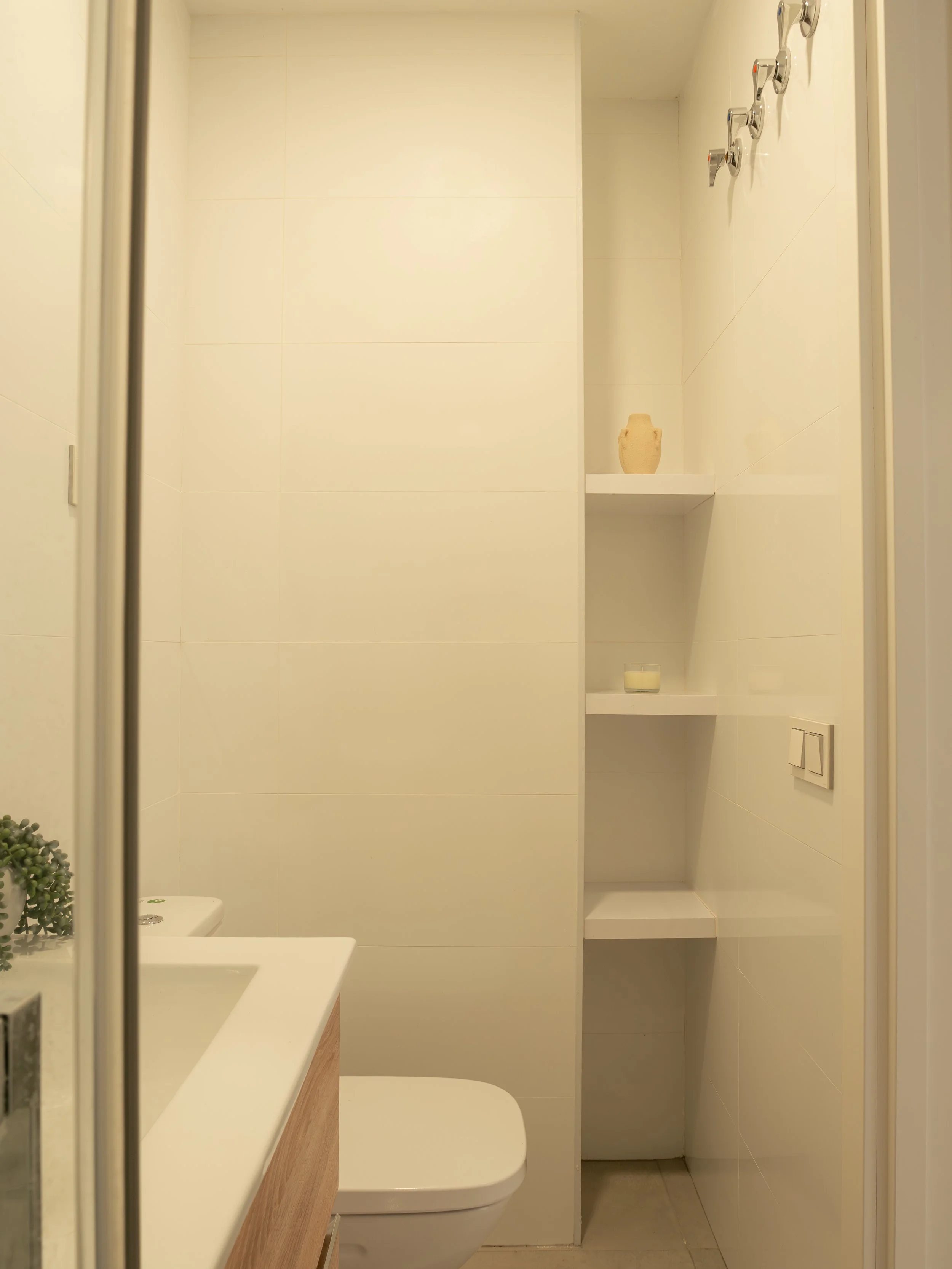 Interior view of a modern bathroom with beige tiles, a white sink, a wooden cabinet, a toilet, and shelves with decorative items, including a vase and a candle.