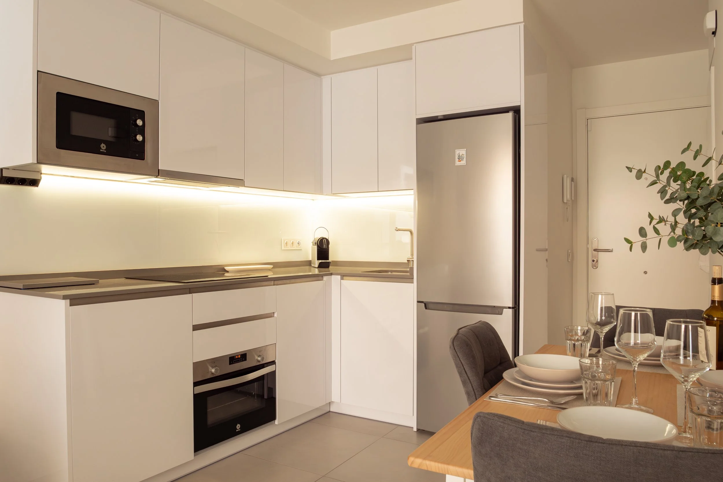 Modern kitchen with white cabinets, stainless steel appliances including a microwave and refrigerator, and a dining area with a table set for a meal featuring glassware and plates.
