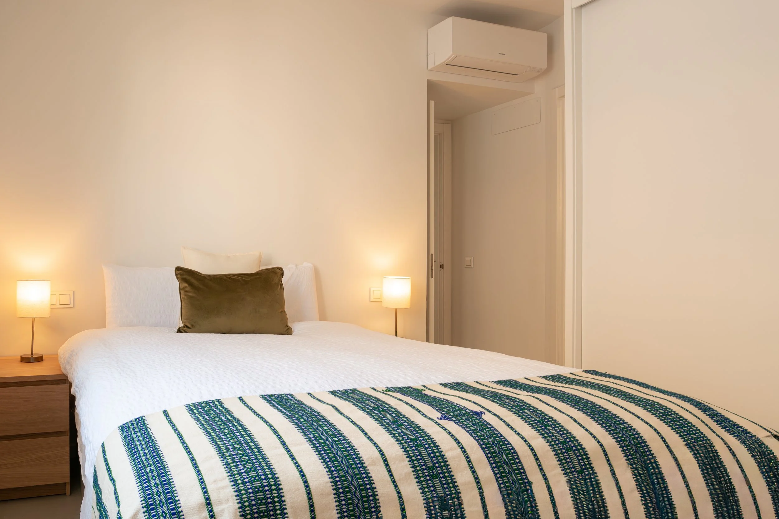 A bedroom with a neatly made bed with white linens and a brown pillow. There are two bedside lamps and a wooden nightstand. The wall is plain, and there is an air conditioning unit above the doorway.