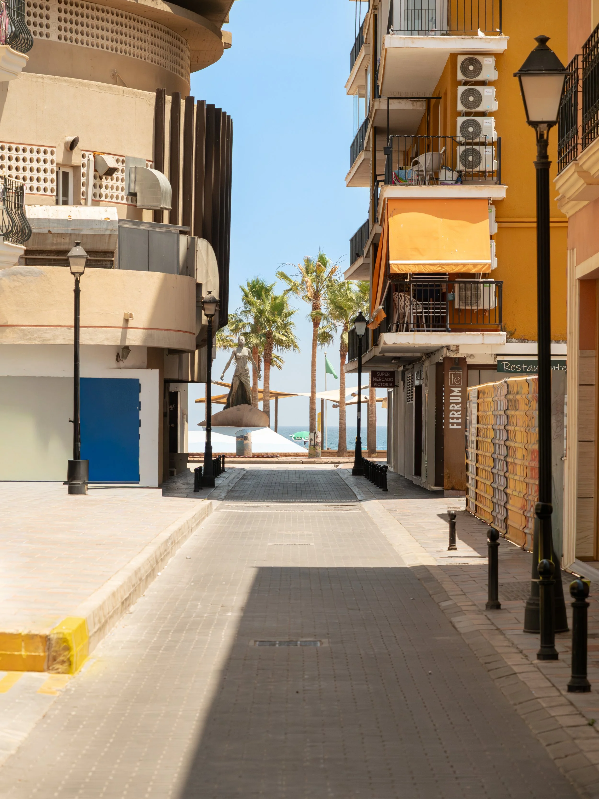 Empty street with colorful buildings, palm trees, and street lamps, leading towards the beach.