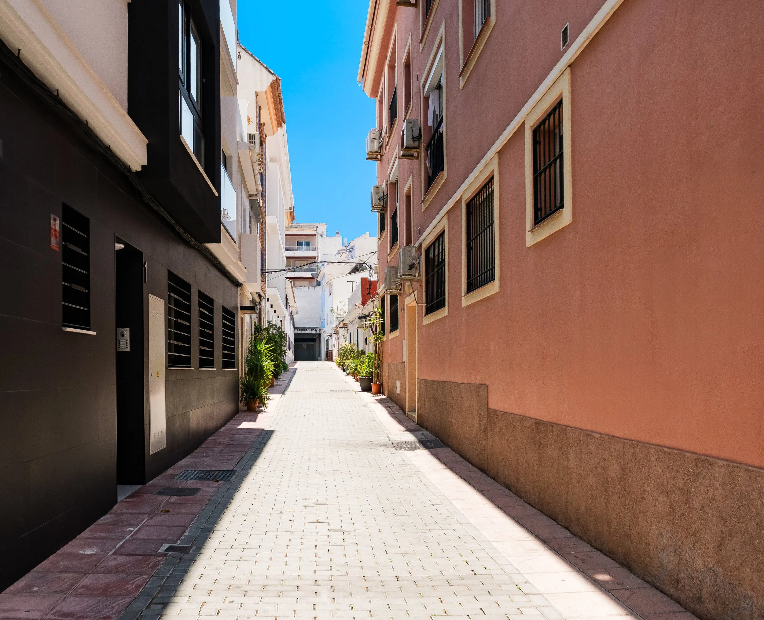 A narrow alleyway lined with apartment buildings on both sides, with potted plants and trees at the base of the buildings. The buildings are painted in various pastel colors, and the clear blue sky is visible above, suggesting a bright, sunny day.