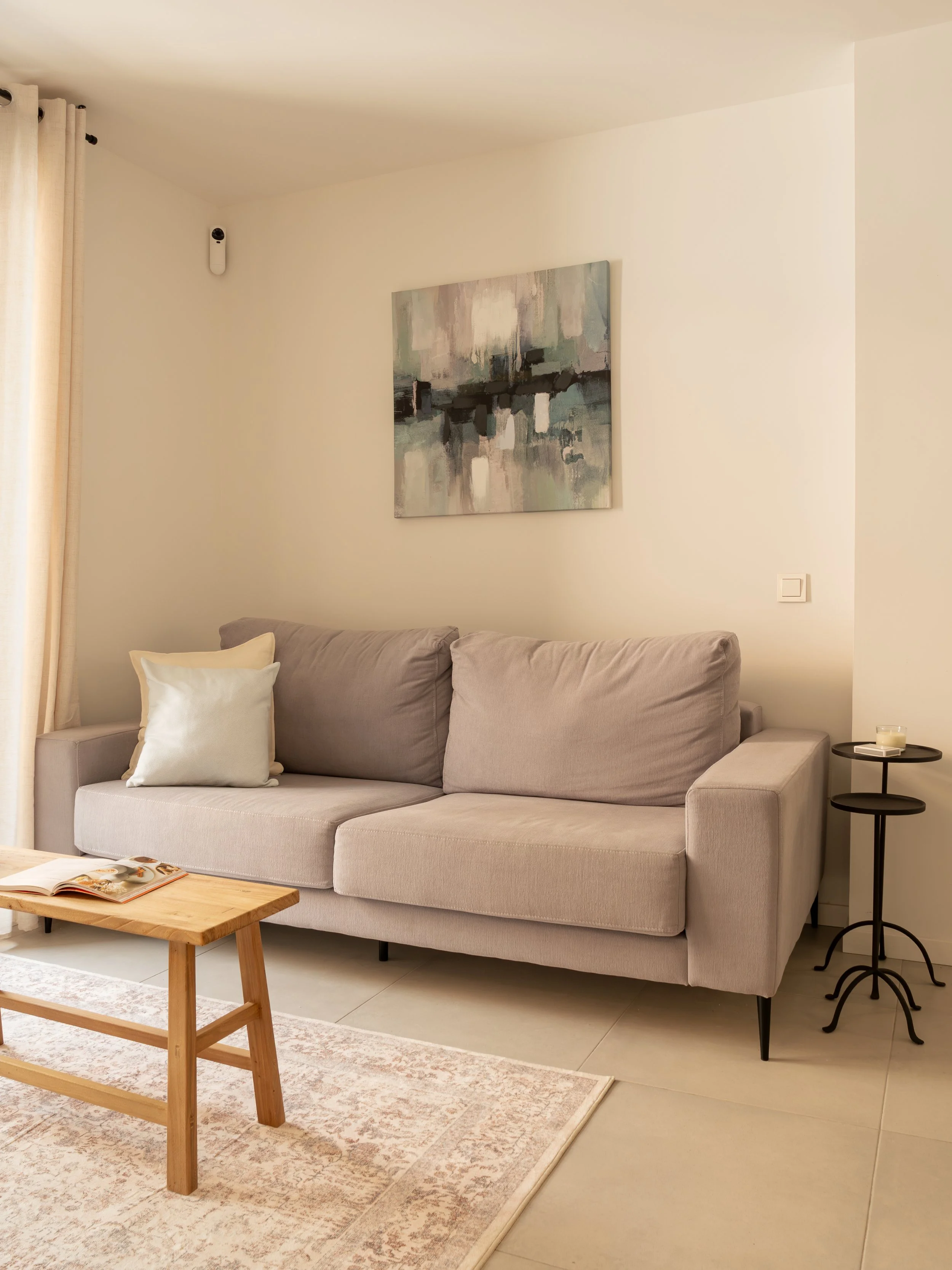 Living room with beige sofa, white pillows, abstract wall art, side table with candle, wooden coffee table, area rug, and curtains.