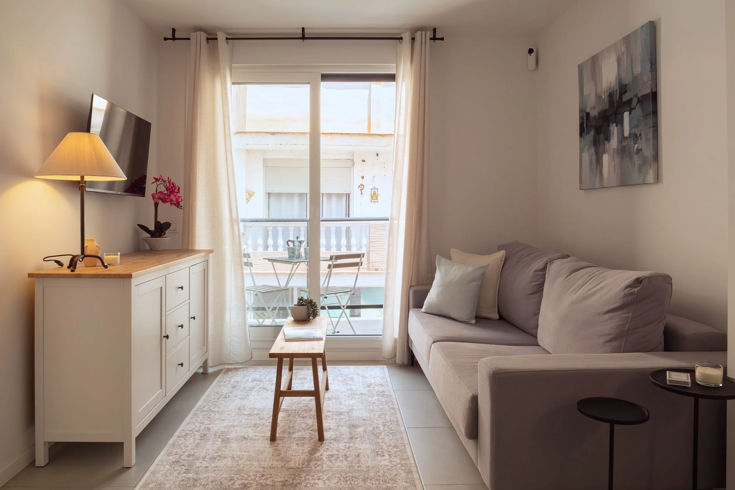 Living room with a beige sofa with pillows, a wooden coffee table, a white sideboard with a pink orchid plant, a lamp, and art on the walls. A sliding glass door with cream curtains leads to a small balcony with a table and two chairs, overlooking an
