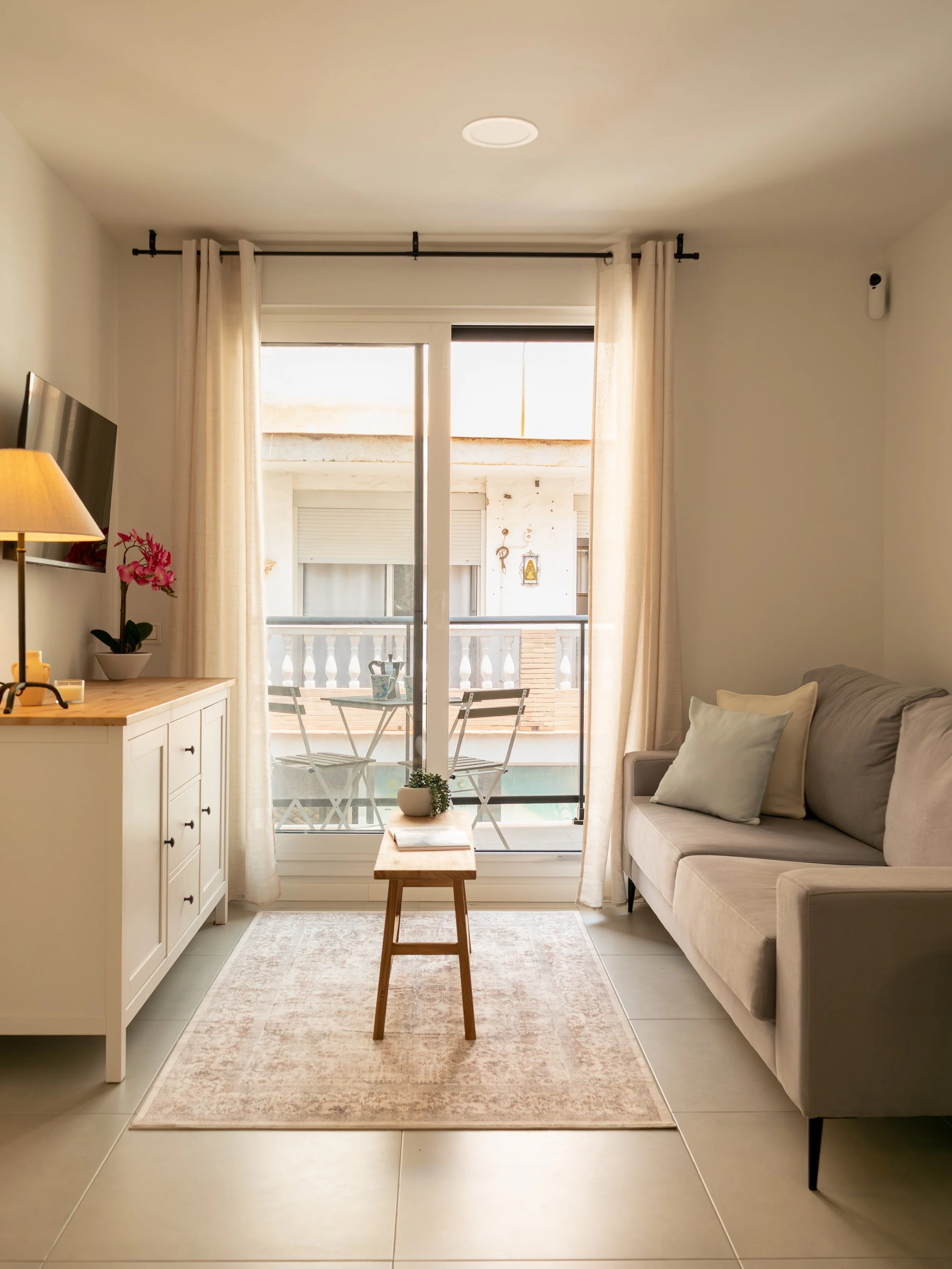 Living room with sliding glass door leading to balcony, beige curtains, gray sofa with pillows, wooden side table with plant, beige rug, and wooden bench.