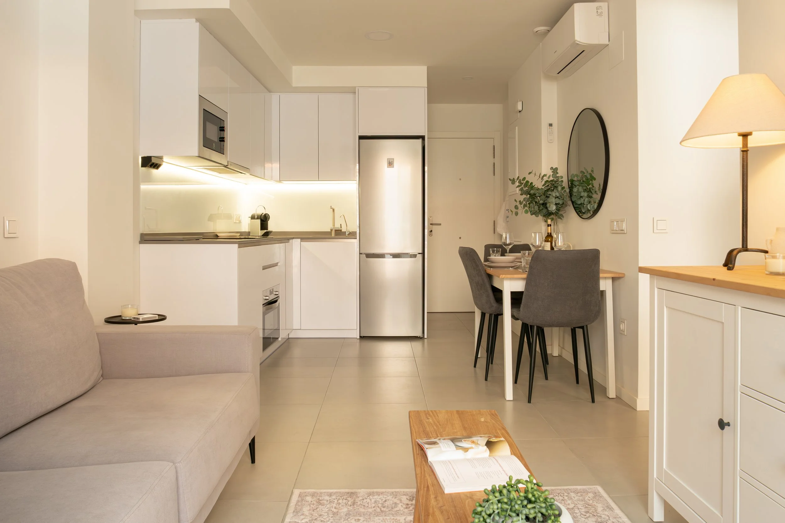 Interior of a modern apartment with a kitchen, dining area, and living room. The kitchen has white cabinets and a stainless steel refrigerator. The dining table is set with glasses and a bottle, and there is a round mirror on the wall. A beige sofa i