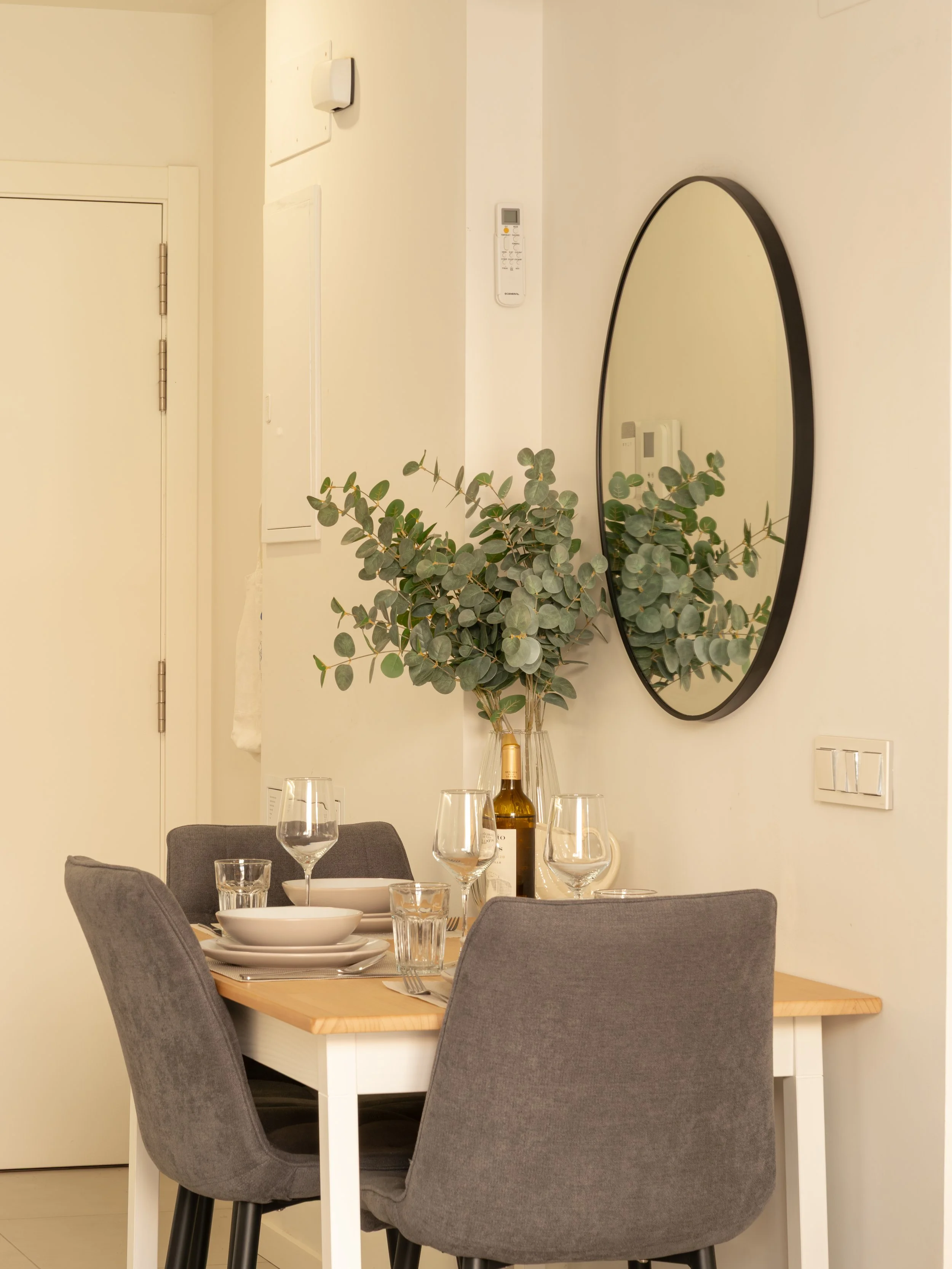 A dining table with grey chairs set with white plates, glasses, and cutlery, and a bottle of wine. A large green plant is in a vase, and a mirror, thermostat, and light switches are on the wall in a bright, modern room.