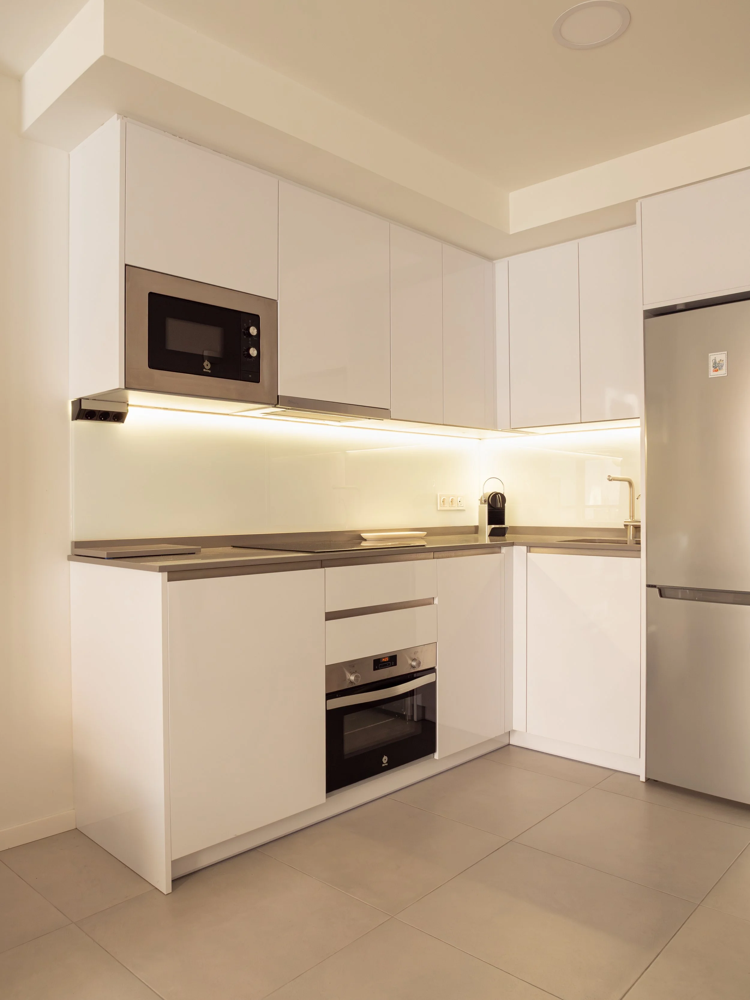 Modern kitchen with white cabinets, stainless steel microwave, oven, and refrigerator, beige tiled floor, and minimalistic design.
