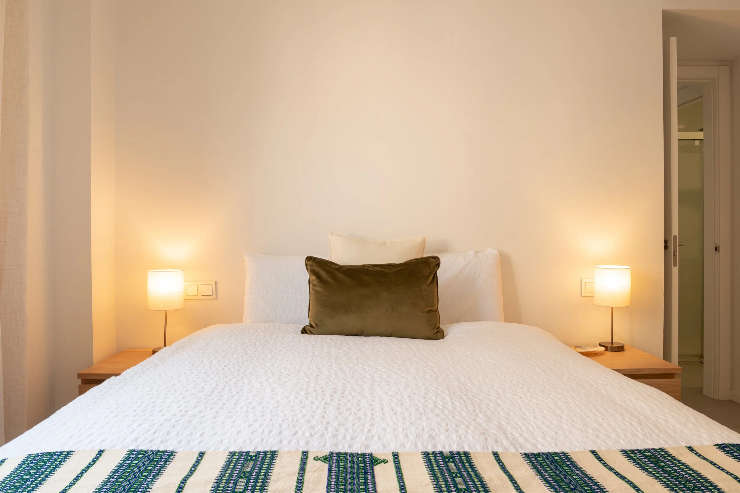 A bedroom with a neatly made bed featuring white bedding and a dark brown pillow, two bedside lamps on wooden nightstands, and a white wall with two electrical outlets behind the lamps. There is a doorway on the right side leading to another room.