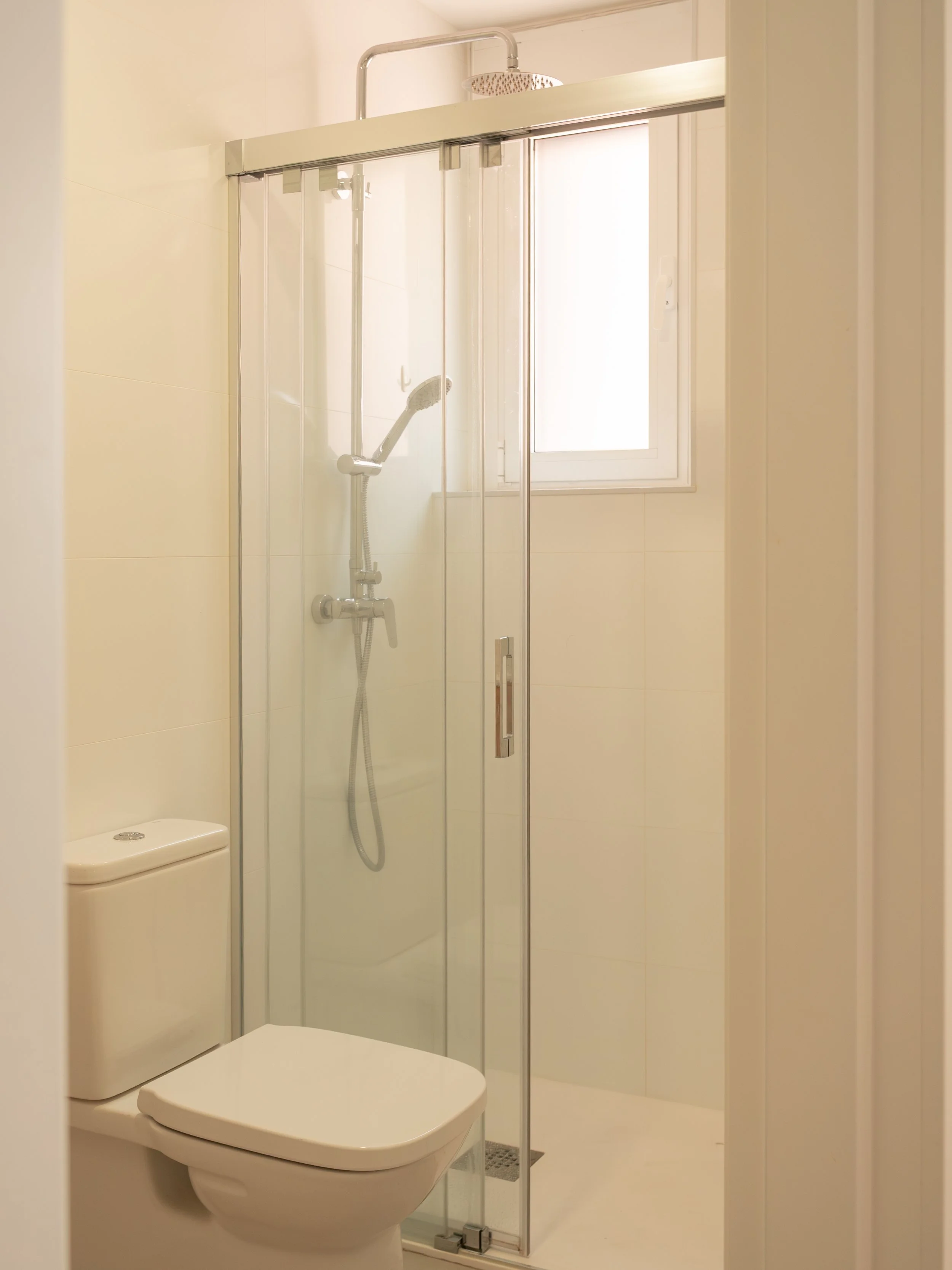 A small modern bathroom with a toilet and a glass shower enclosure. The shower has a showerhead and a hand-held shower wand, and there is a window allowing natural light into the space.