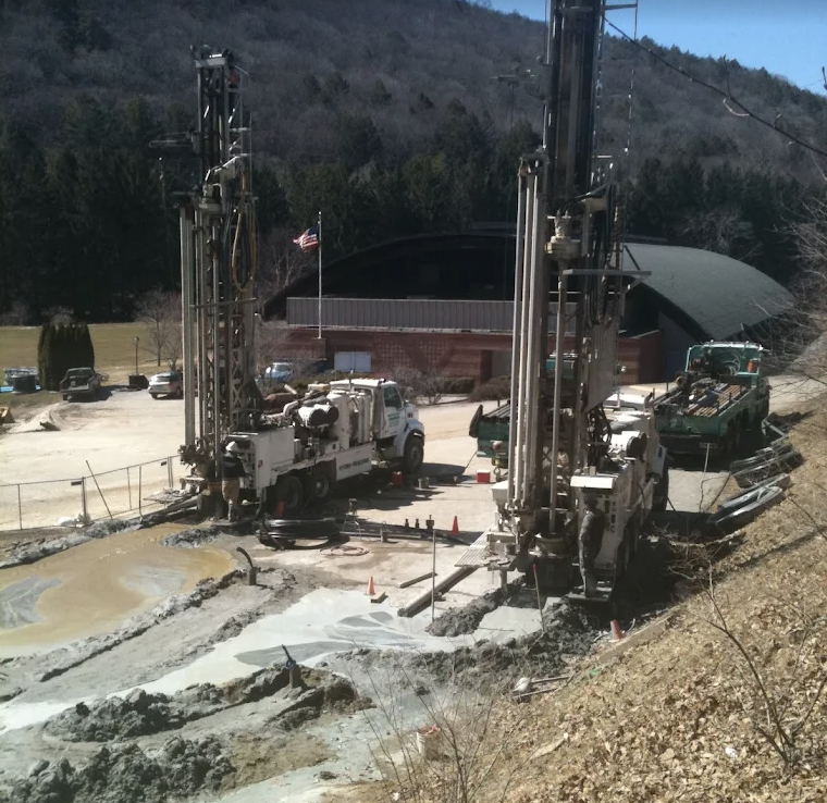 Construction site with two large water well drilling machines and a pickup truck, with a building and trees in the background.