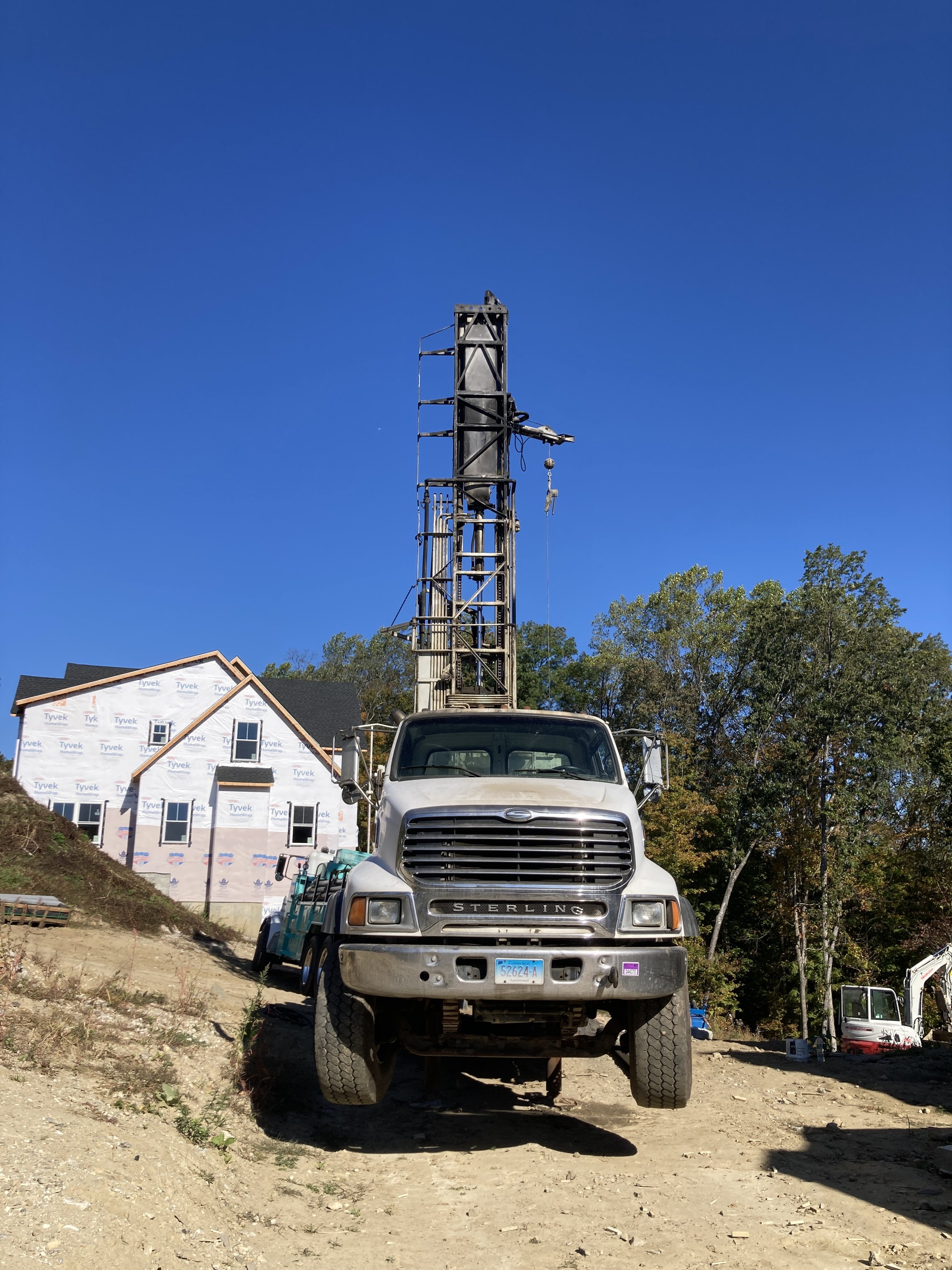 A construction site featuring Wragg Well Drilling's truck with a tall drilling rig on a clear, sunny day. In the background, there is a house under construction with white weather-resistant sheathing and a black roof, surrounded by trees.