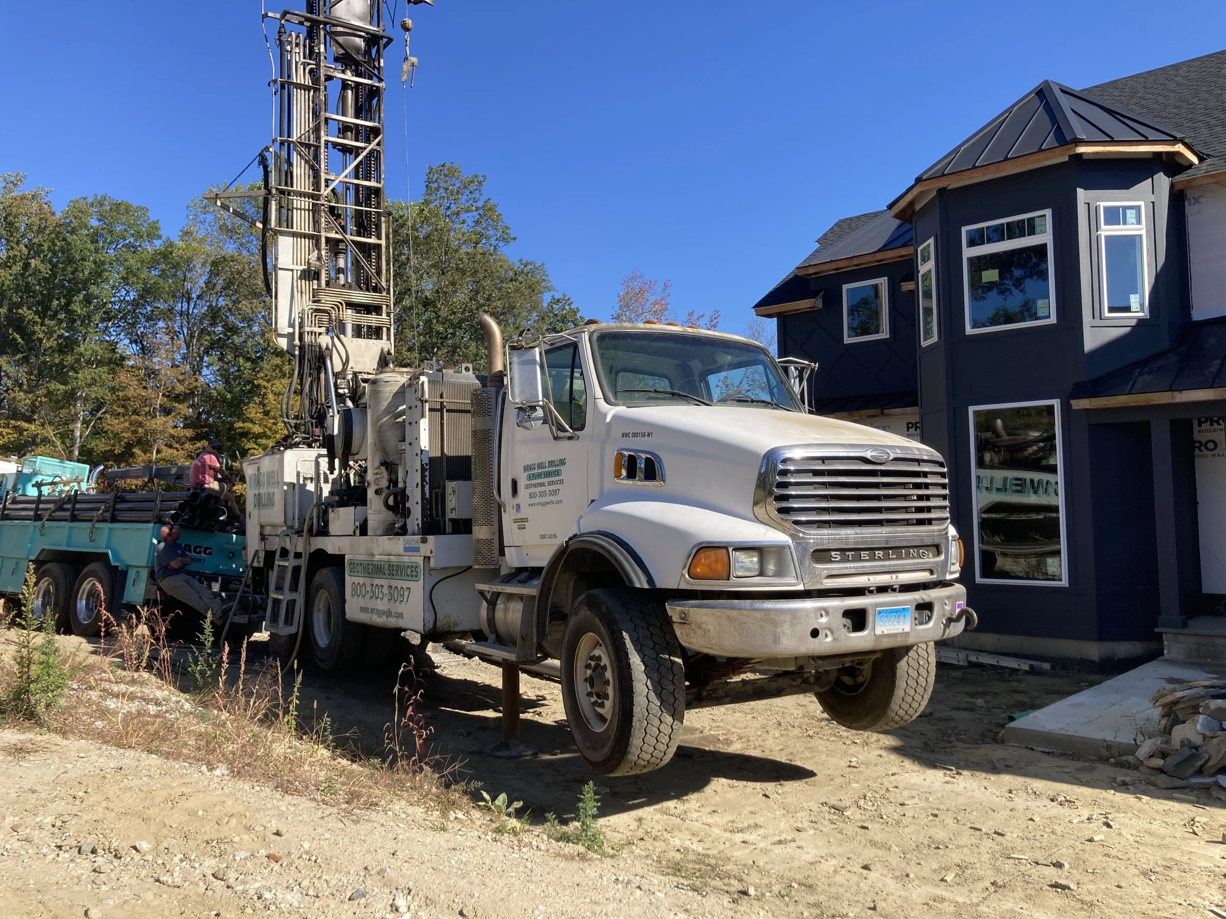 Construction site with  Wragg Well drilling's truck parked next to a dark blue house under construction. The truck has a tall drilling apparatus and workers nearby, with trees in the background and a clear blue sky.
