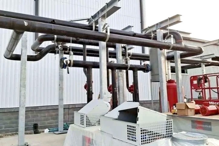 Industrial piping system with black pipes, valves, and red fire suppression equipment outside a building.