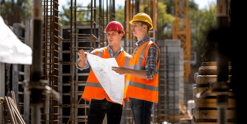 Two construction workers in orange safety vests and helmets discussing blueprints at a construction site with steel reinforcement bars and building materials around.