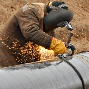 Worker wearing a welding helmet and gloves, welding a large metal pipe, with sparks flying.
