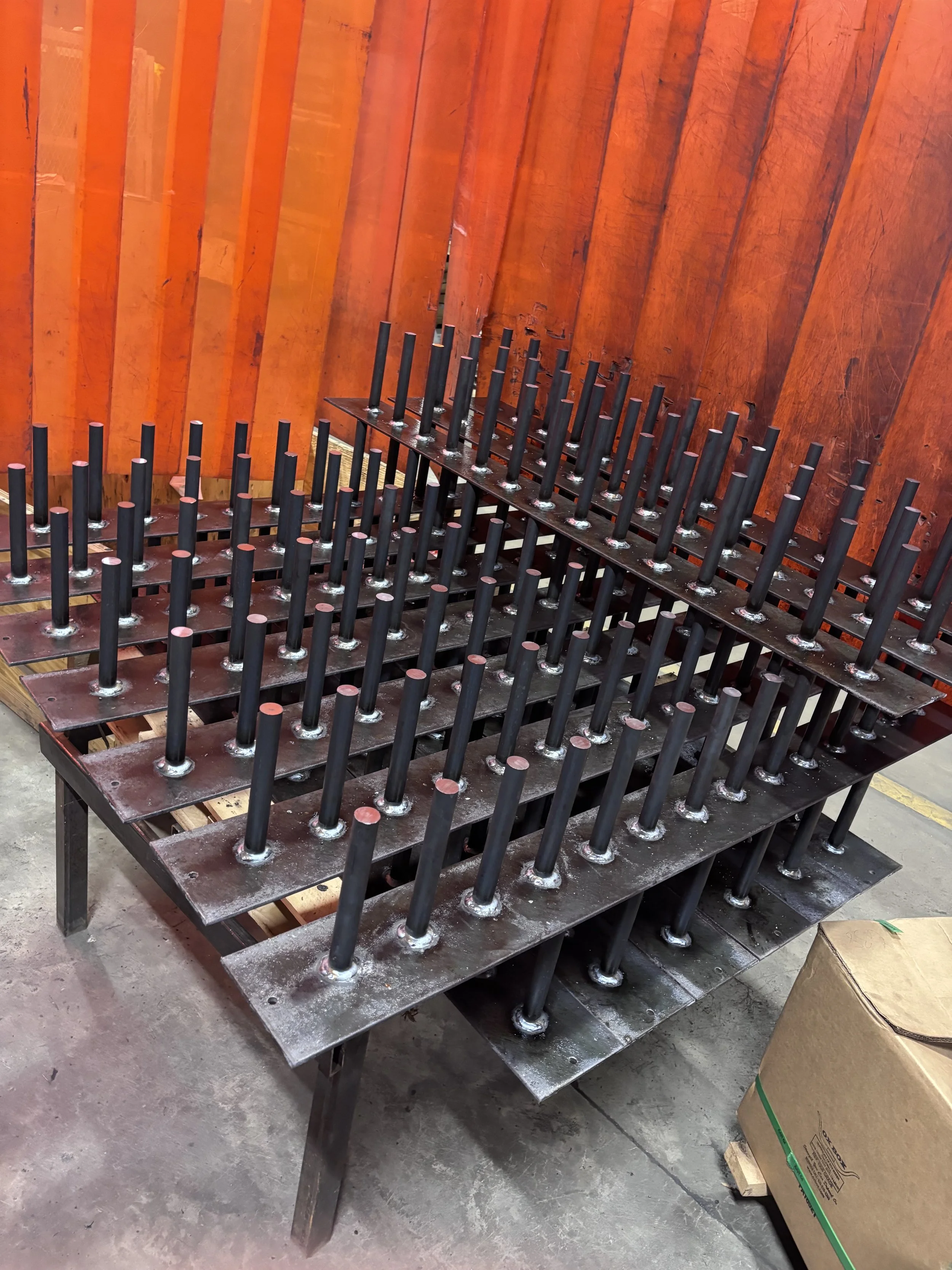 A metal table with multiple vertical black rods welded onto flat metal pieces, arranged in a grid pattern, placed against orange wooden panels in an industrial setting.