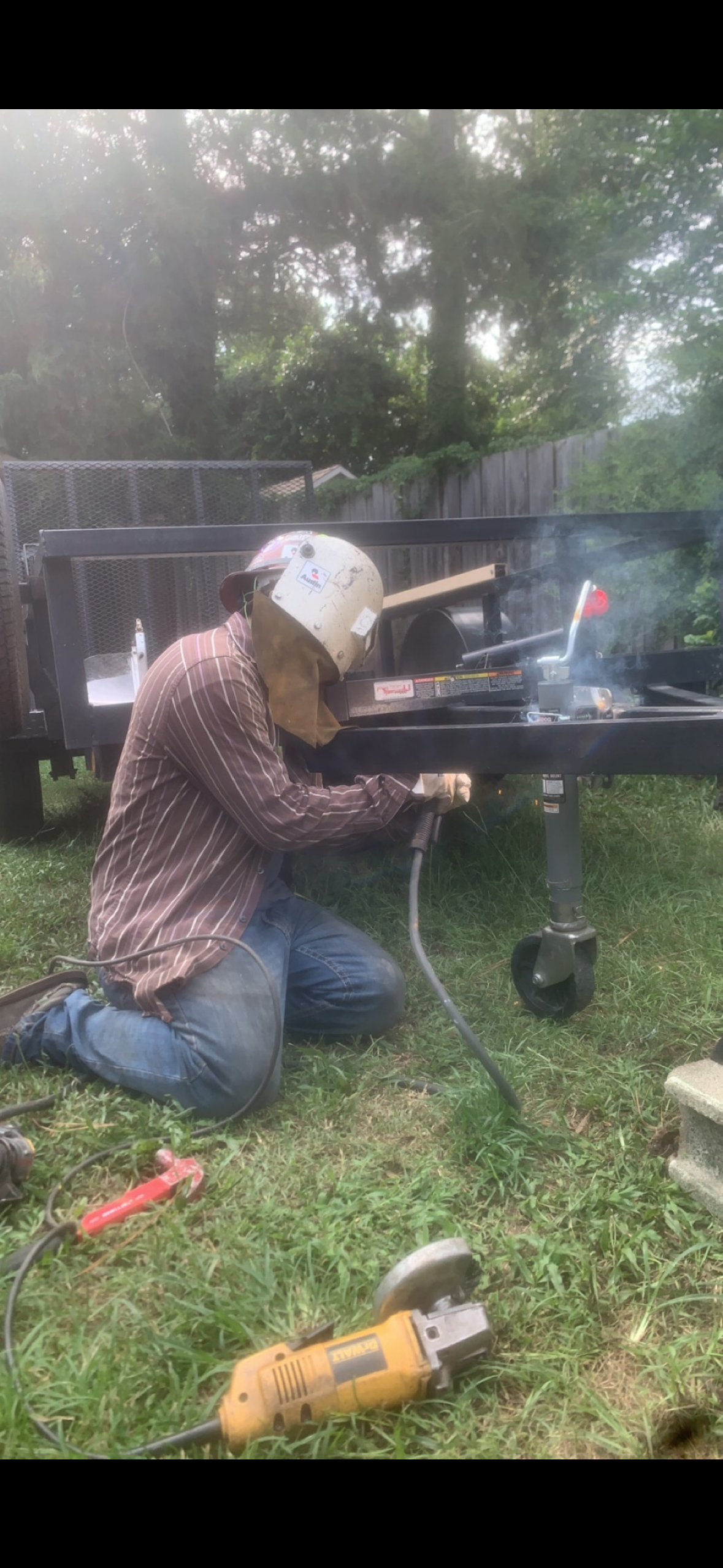 A person welding outdoors on a black trailer, kneeling on grass, wearing a helmet and protective gear, with welding equipment and tools around.