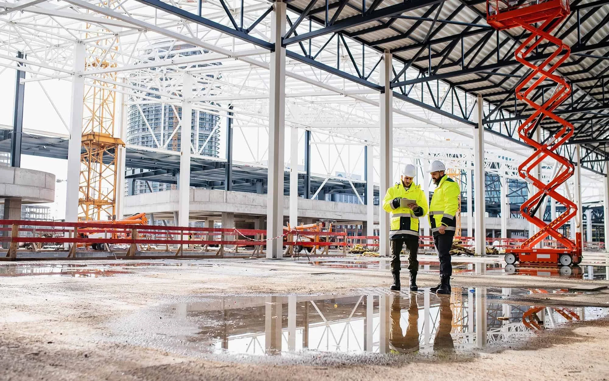 Two construction workers in yellow safety jackets and white helmets standing on muddy ground, looking at a tablet at a building site with steel framework and a scissor lift in the background.