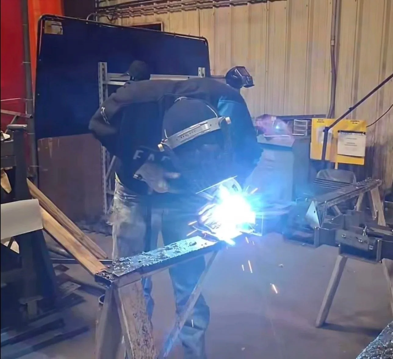 Welder in a workshop continuously welding metal on a workbench, with sparks flying and bright blue welding light illuminating the scene.