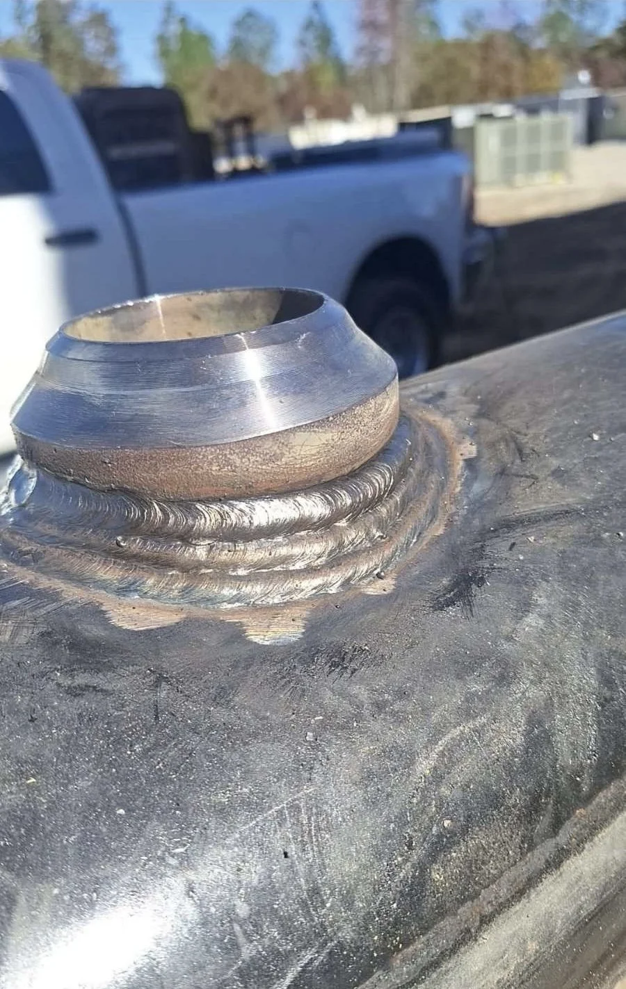 Close-up of a welded metal fitting on a large metal surface, with a pickup truck and trees in the background.