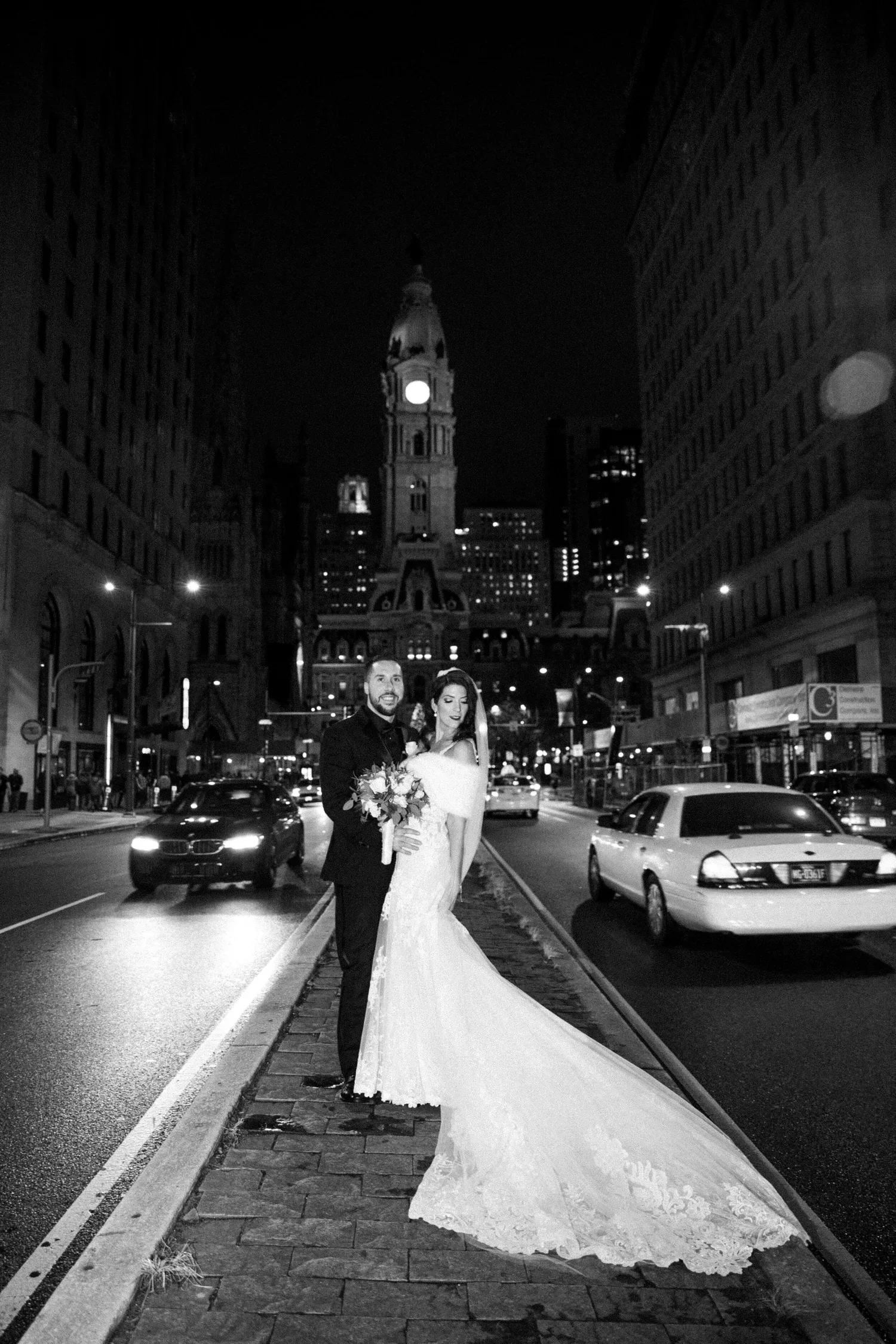A black and white photo of a newlywed couple standing on a city street at night, with a historic clock tower in the background. The bride is holding a bouquet and wearing a long, elegant wedding dress with a train, while the groom is in a dark suit.