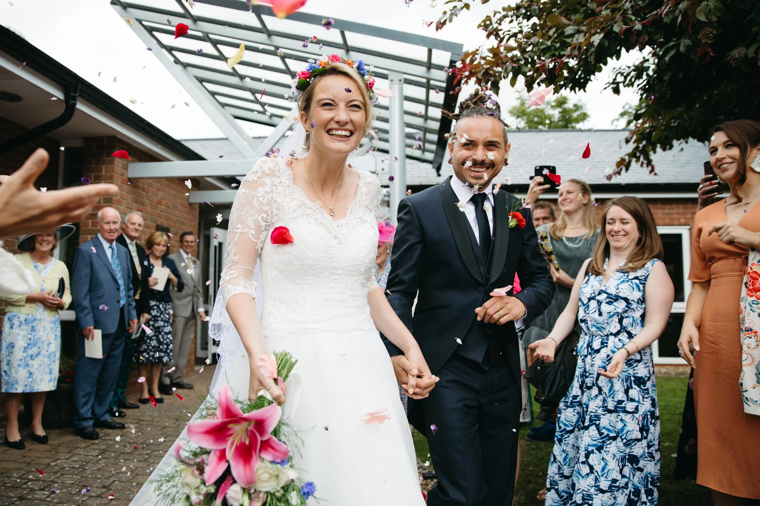 A newlywed couple, bride in a white wedding dress holding a bouquet and groom in a black tux, walking hand in hand surrounded by guests celebrating and throwing flower petals outside a building.