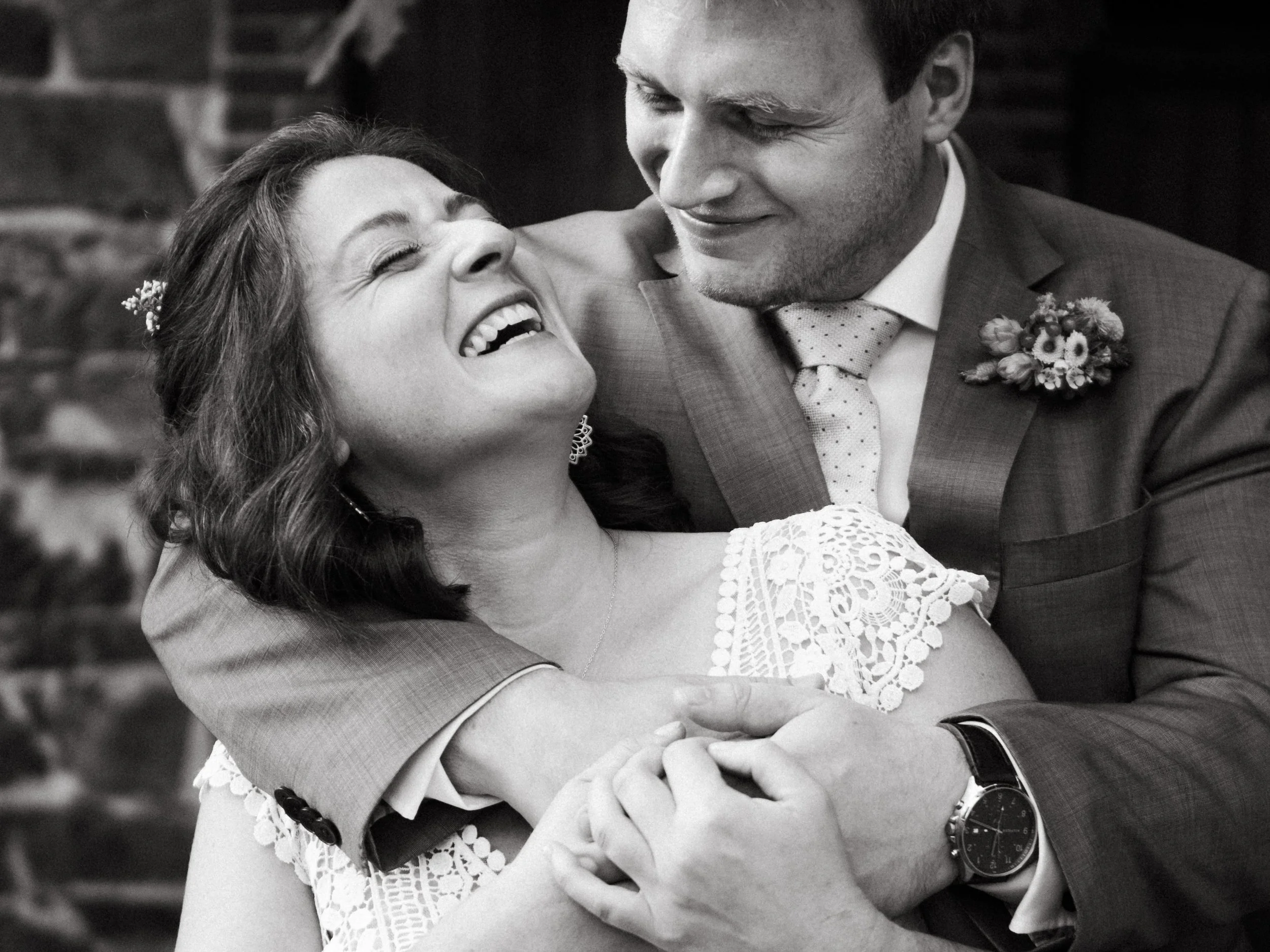 A joyful woman and a man in a suit embrace during a wedding. The woman is smiling with her eyes closed, and the man is looking at her with a smile. The woman wears a lace dress and earrings, while the man wears a suit, tie, boutonniere, and a watch.