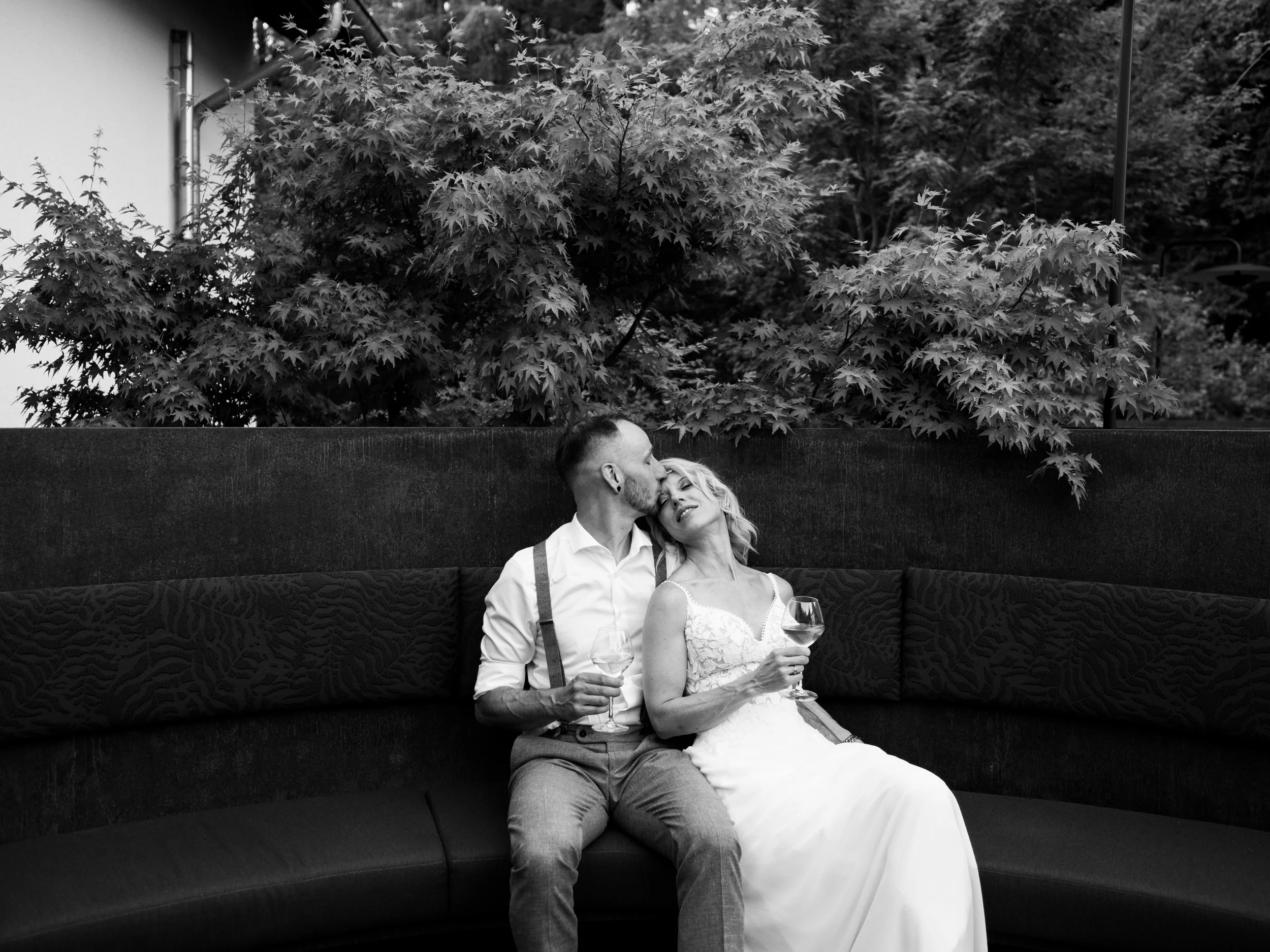A black and white photo of a couple sitting on a curved outdoor bench, with trees in the background. The woman is in a wedding dress holding a glass of wine, and the man is in a shirt and suspenders, also holding a glass of wine. The man is kissing t