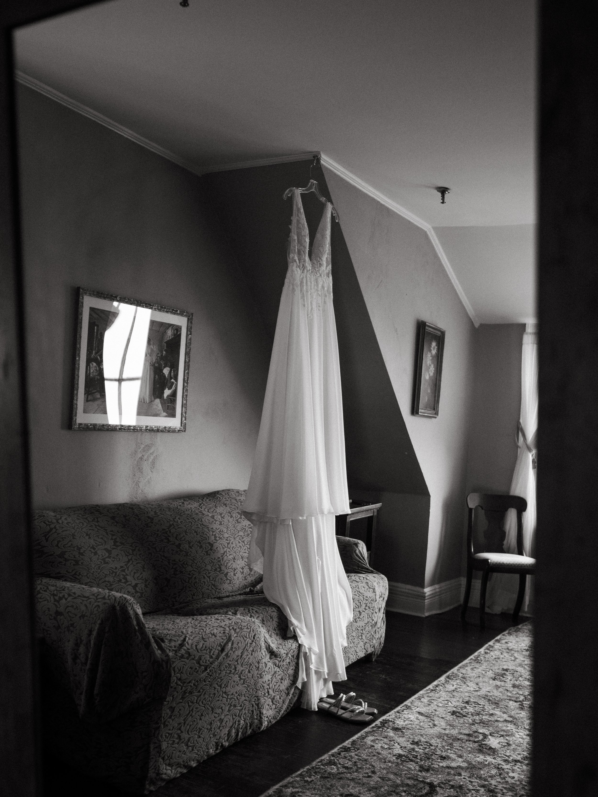 A wedding dress hanging from the ceiling in a living room, with a patterned sofa, framed pictures on the wall, a chair, and a curtain in the background.