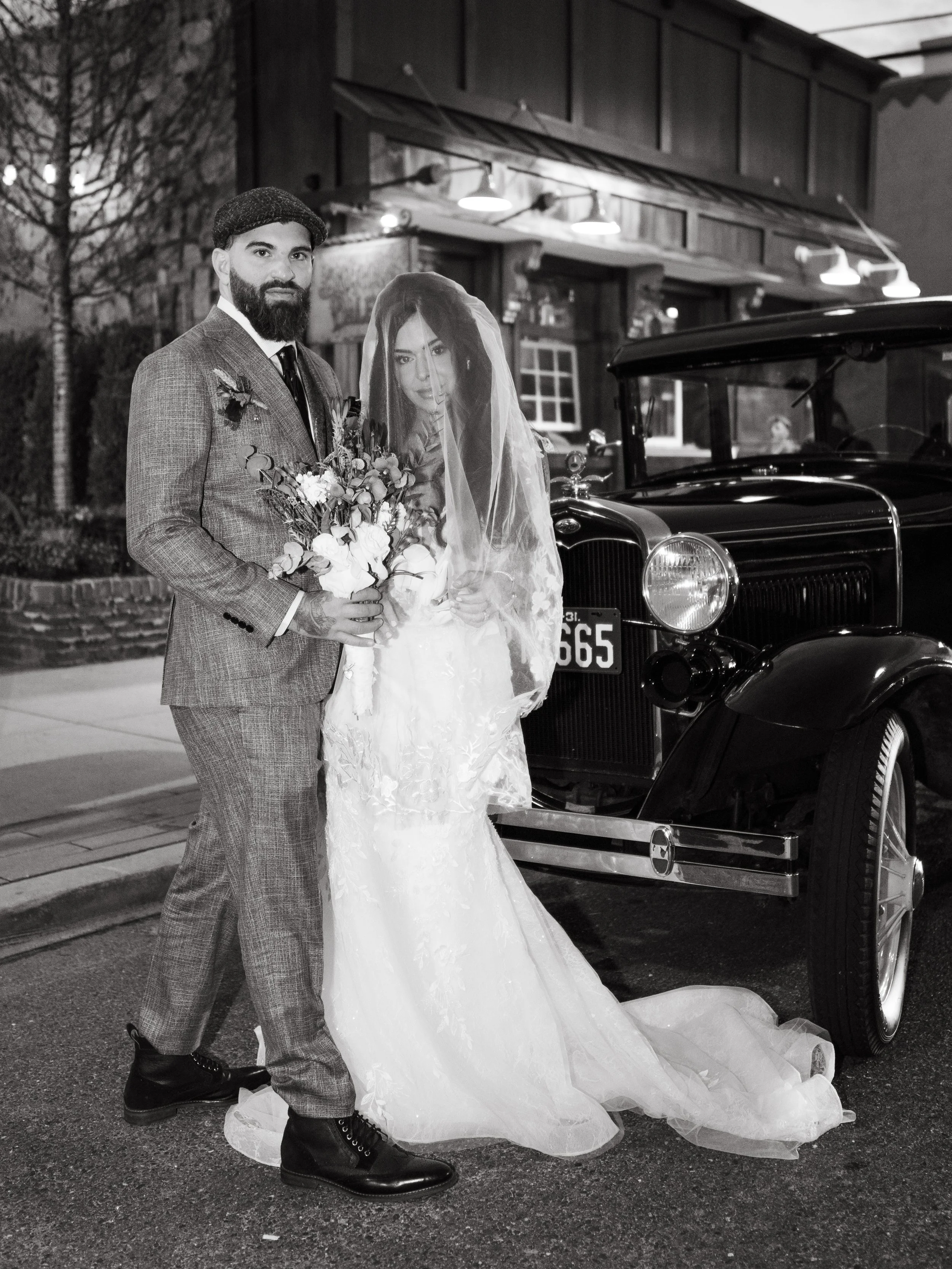 Black and white photo of a bride and groom standing beside a vintage car. The groom is dressed in a suit with a tie and holding a bouquet. The bride wears a wedding dress and veil. They are outdoors, possibly on a city street at night.