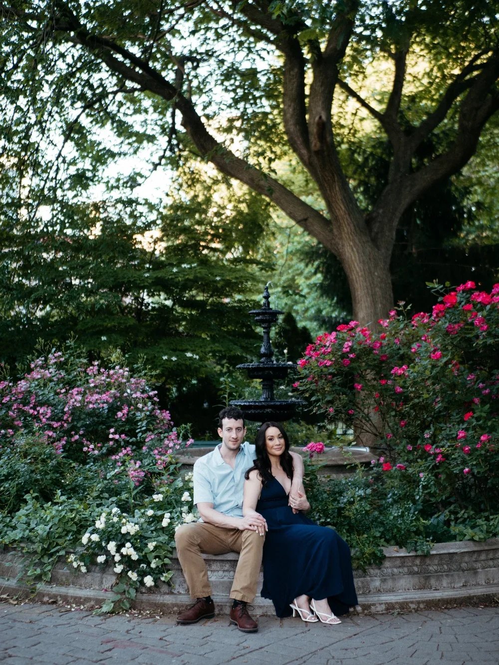 A couple sits together on a stone bench in a garden with pink and white flowers, green foliage, a fountain, and a large tree, during sunset.