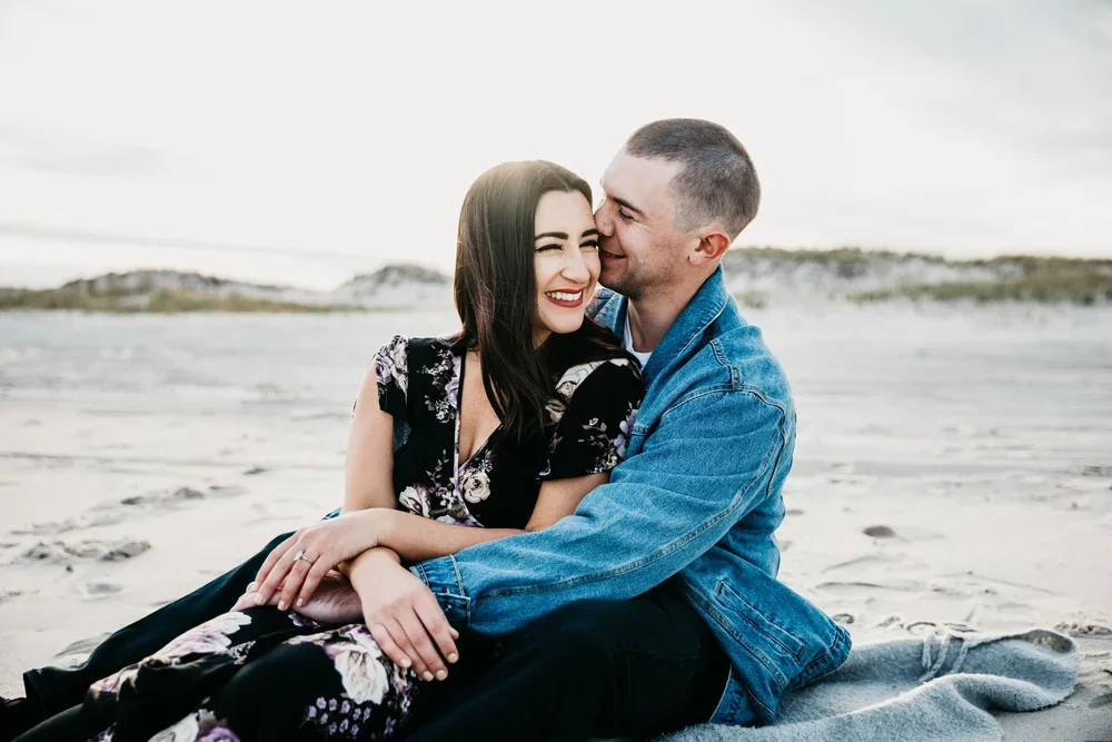 A happy couple sitting on the beach, embracing and smiling, with a sandy shoreline and dunes in the background.