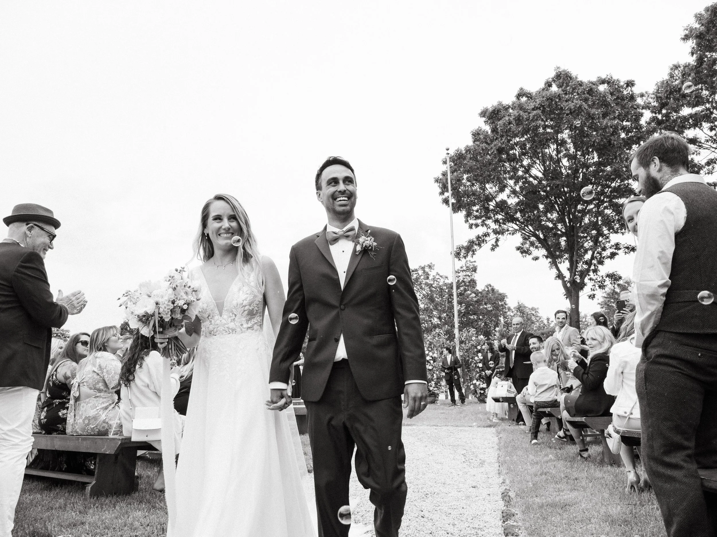 Black and white photo of a newlywed couple walking hand in hand outdoors, surrounded by seated guests and standing onlookers, with trees in the background, celebrating at their wedding.