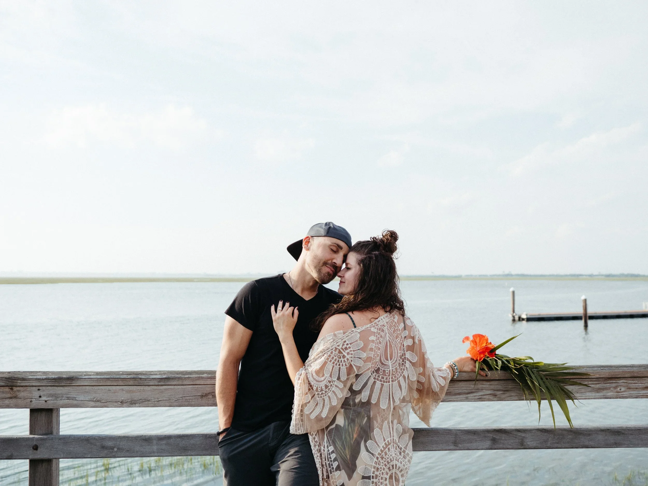 Couple standing close together on a wooden pier by a body of water, with the woman holding a bouquet of red and orange flowers.
